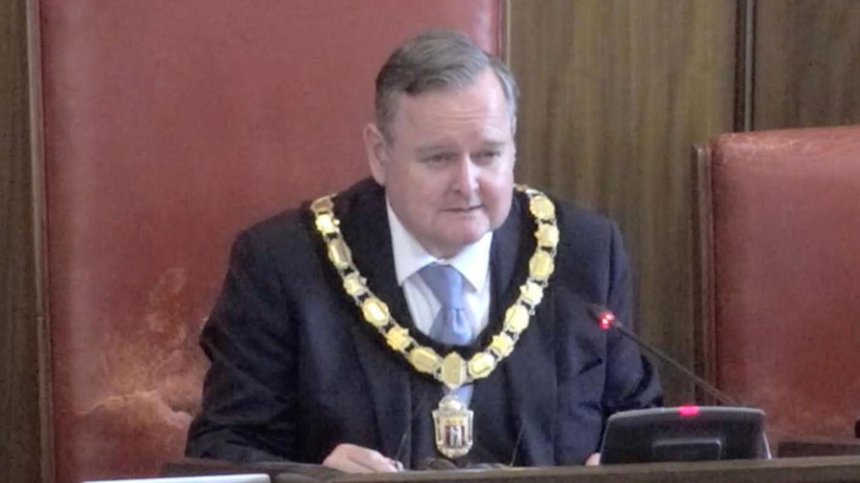 Warwickshire County Council chairman and Reform councillor Edward Harris pictured in the red leather Chairman's seat in the full council chamber in Shire Hall. He is wearing a gold ornate chain, blue suit and tie.