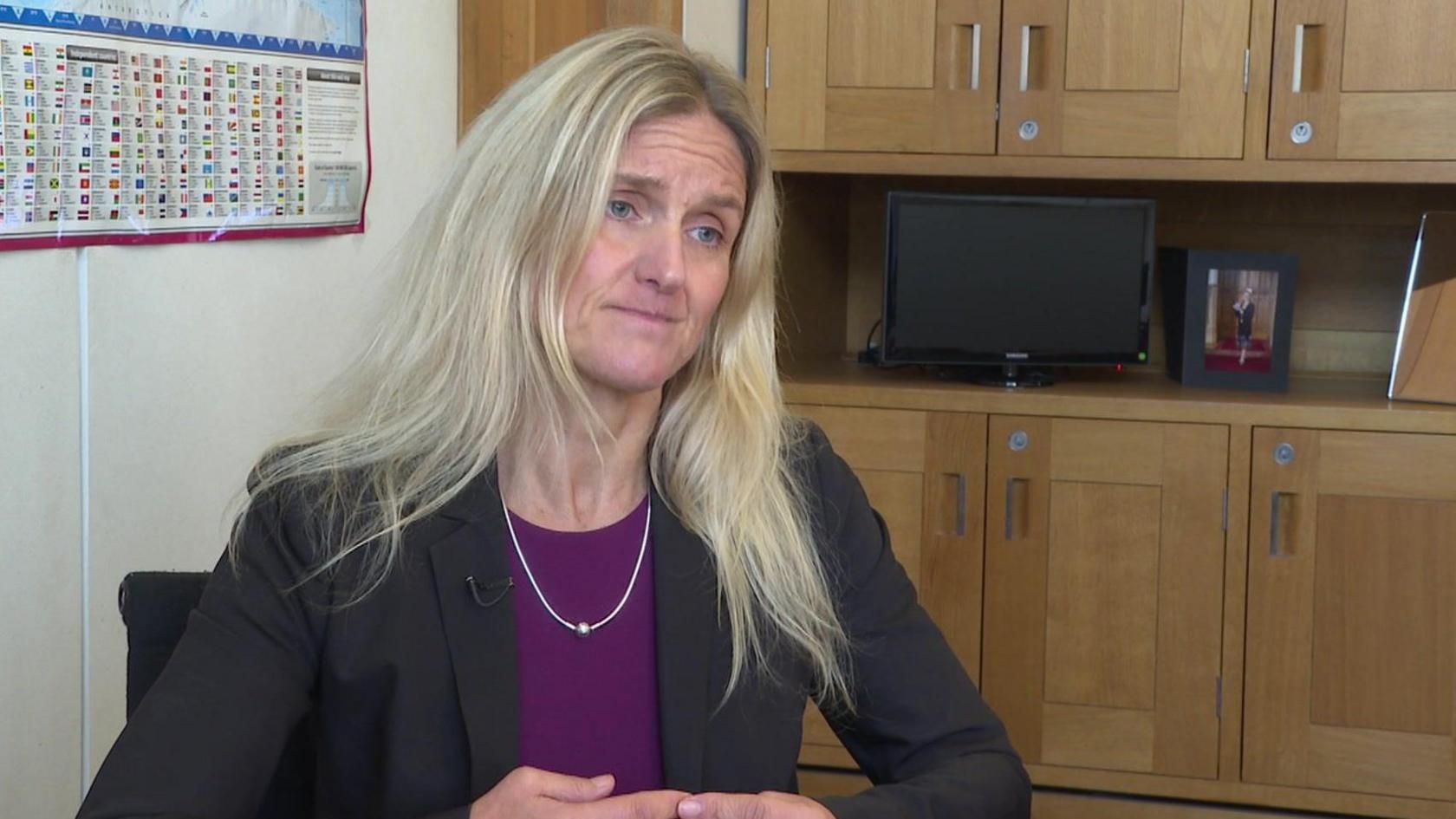 MP Kim Leadbeater sits in her office. She has shoulder-length blonde hair and is wearing a black jacket and purple top. Behind her is an oak cabinet with a small TV on it.