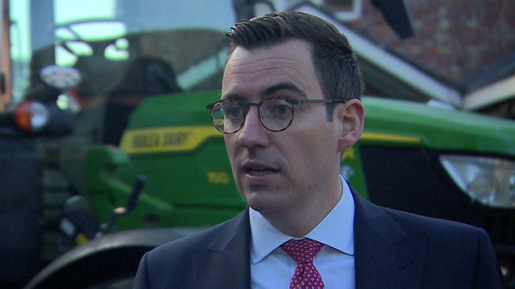 Dan Price, in a suit with a pink tie over a white shirt and wearing glasses, stands in front of a green tractor