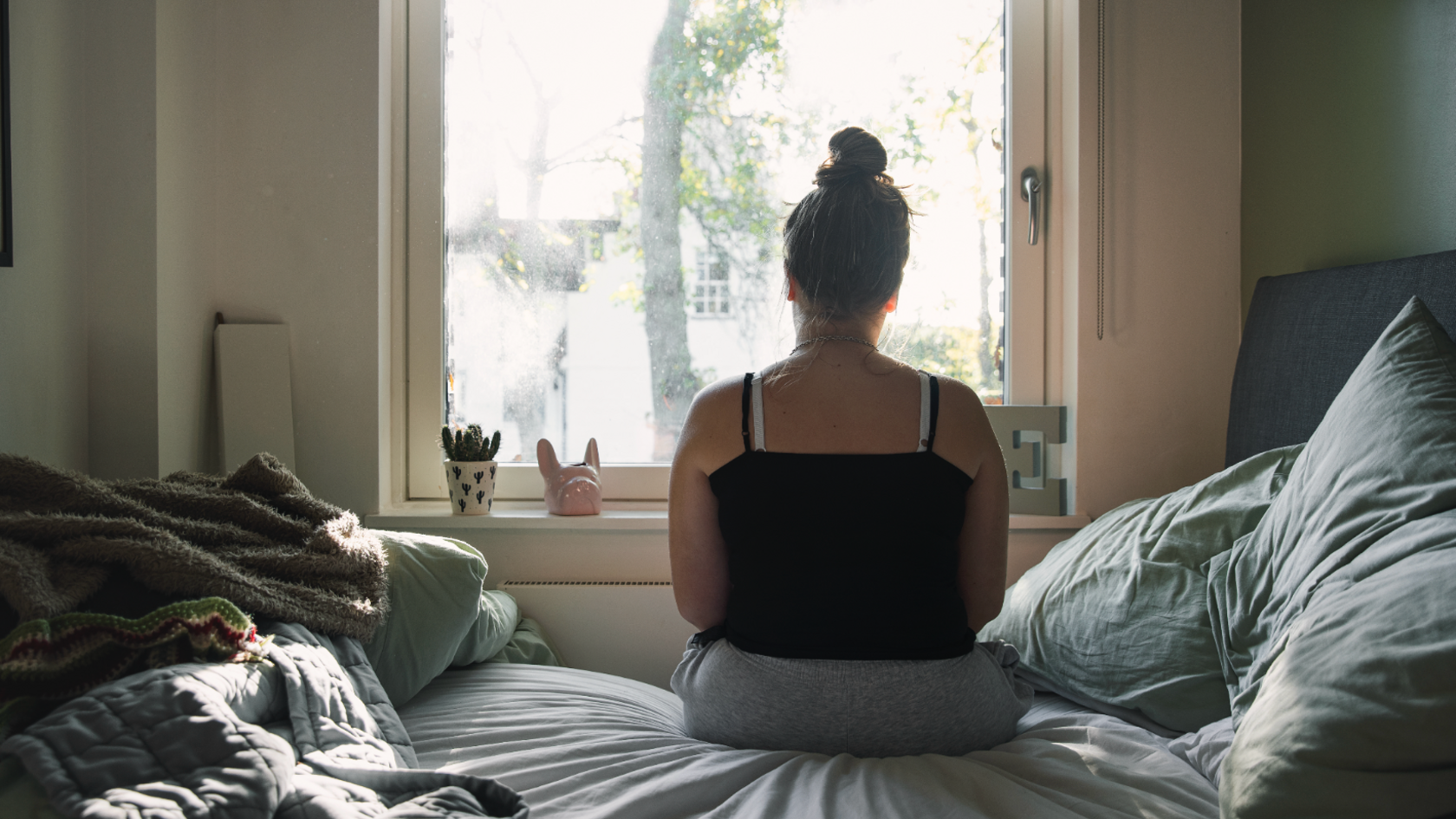 young woman sitting in bedroom facing a window with her back to the camera