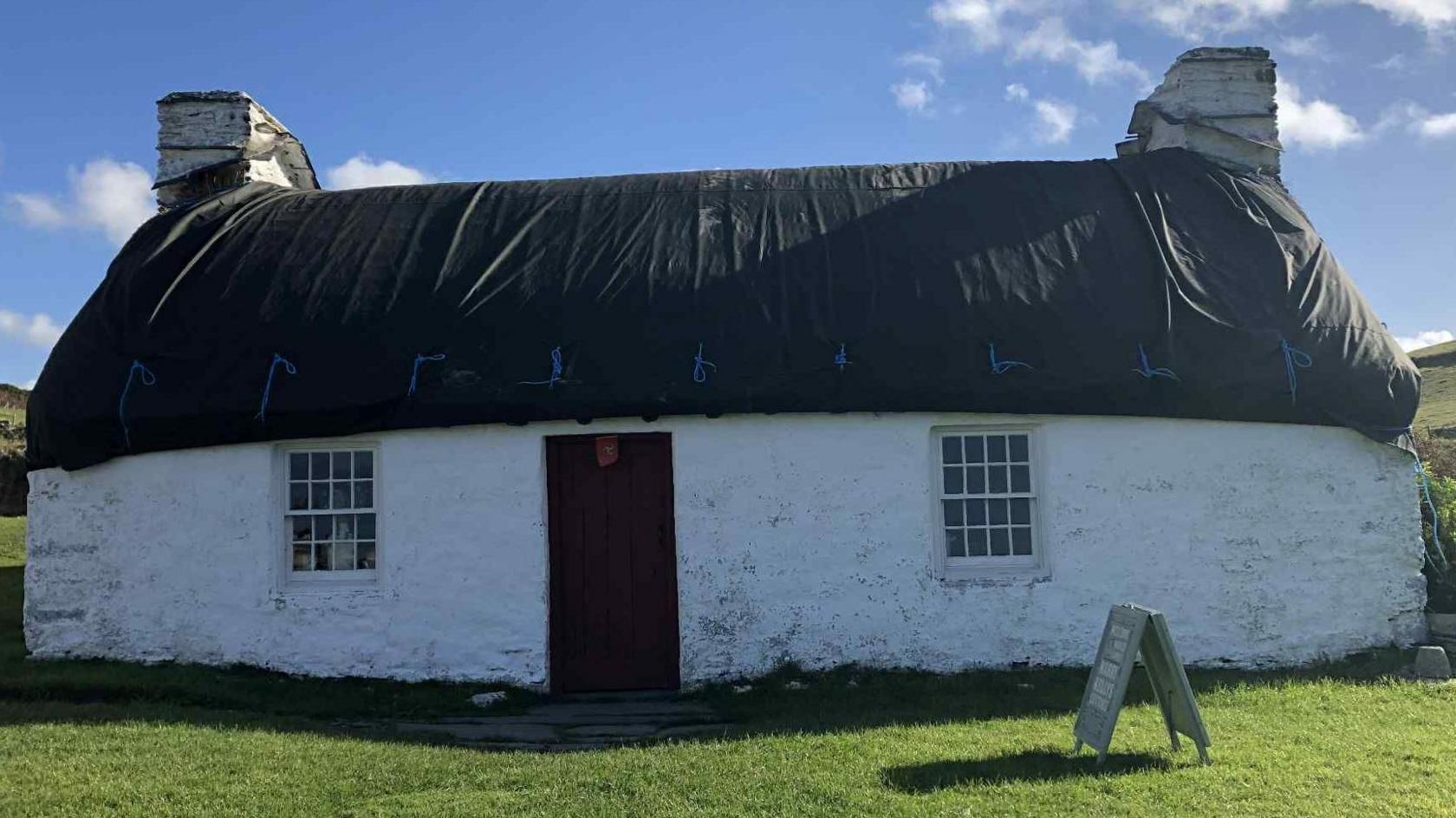 A white Manx stone cottage with a chimney stack at each end, which has its thatch roof covered in a black tarpaulin.