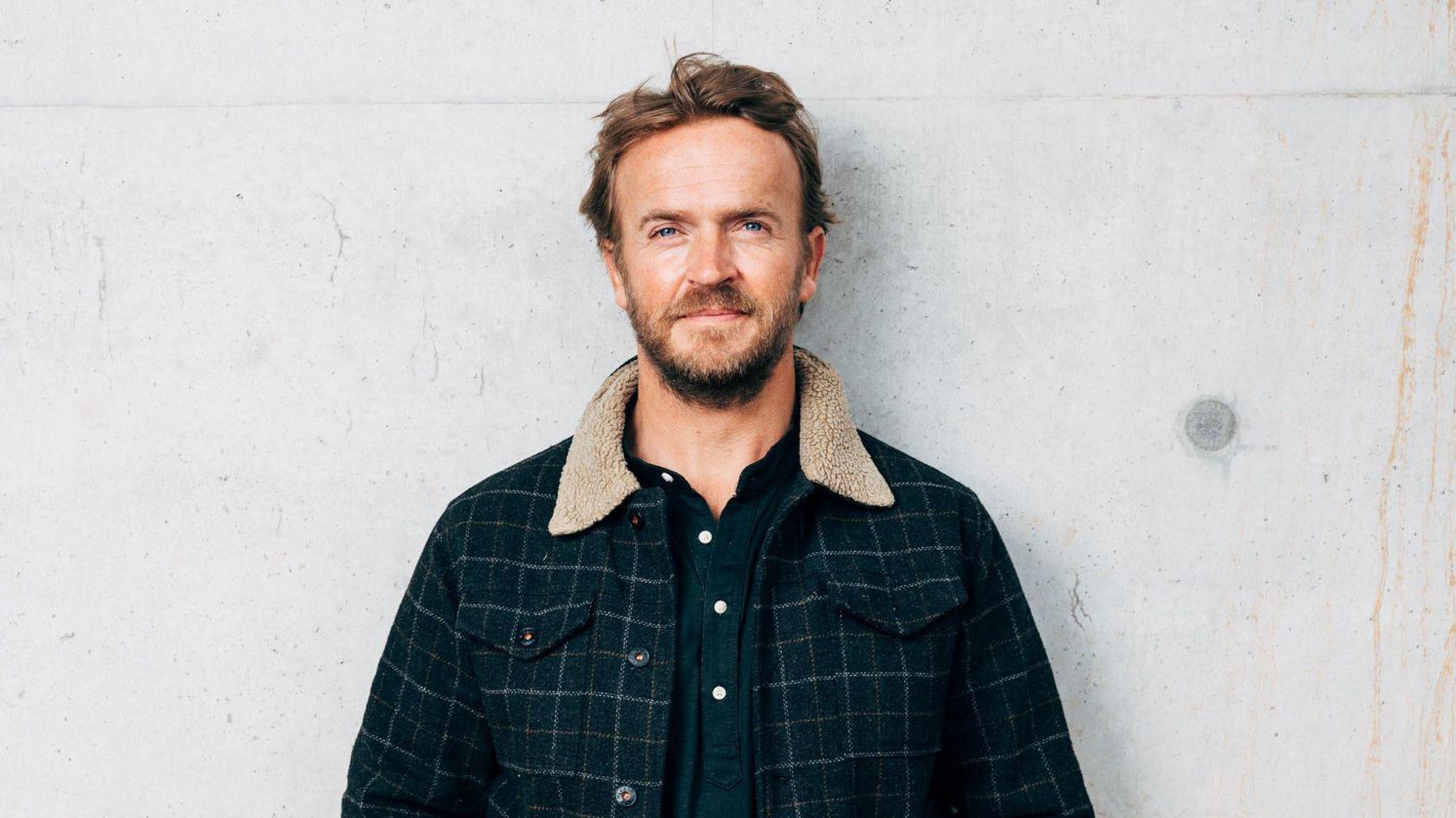 Nick Hounsfield standing against a white concrete wall. He is wearing a dark buttoned shirt and a checked patterned jacket with a fluffy collar. He has blue eyes, short brown hair, a full brown beard, and is smiling at the camera.