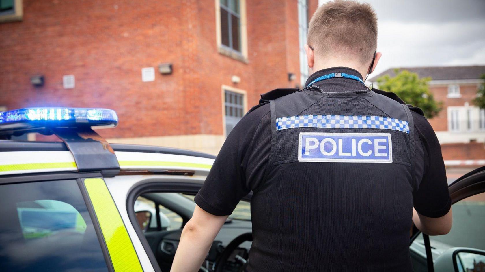 A police officer in a black uniform enters a police car which has its blue lights on.