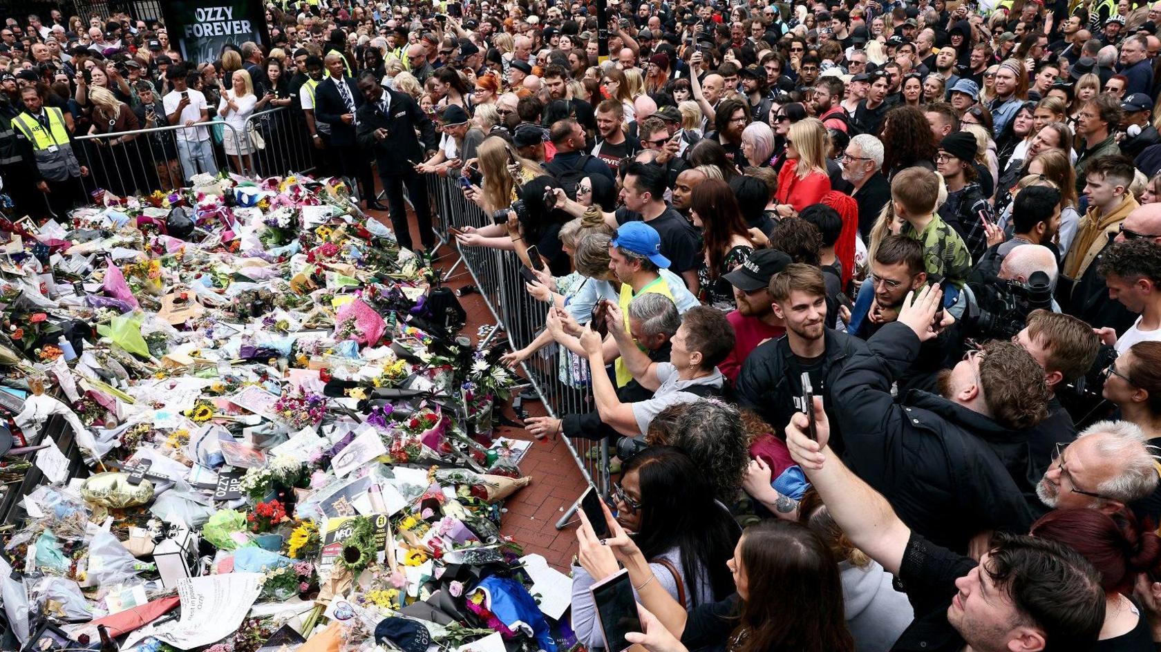 Thousands of people are standing in a crowd in front of a pile of floral and paper tributes. A metal fence surrounds the pile of gifts. There is a board within the crowd that reads "ozzy forever"