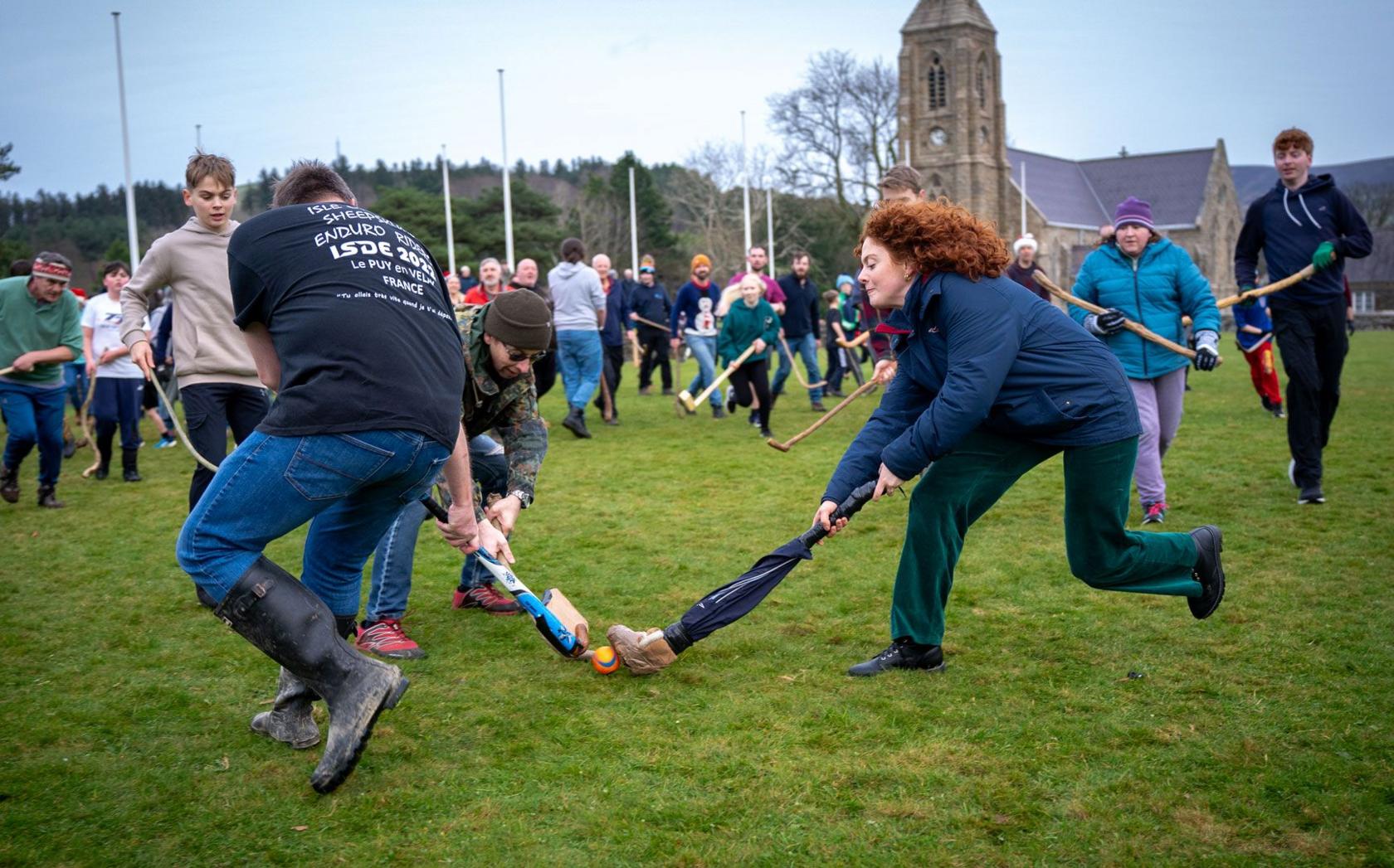 A woman with a homemade stick competes with two other players for the ball. The Royal Chapel of St John's can be seen in the background.