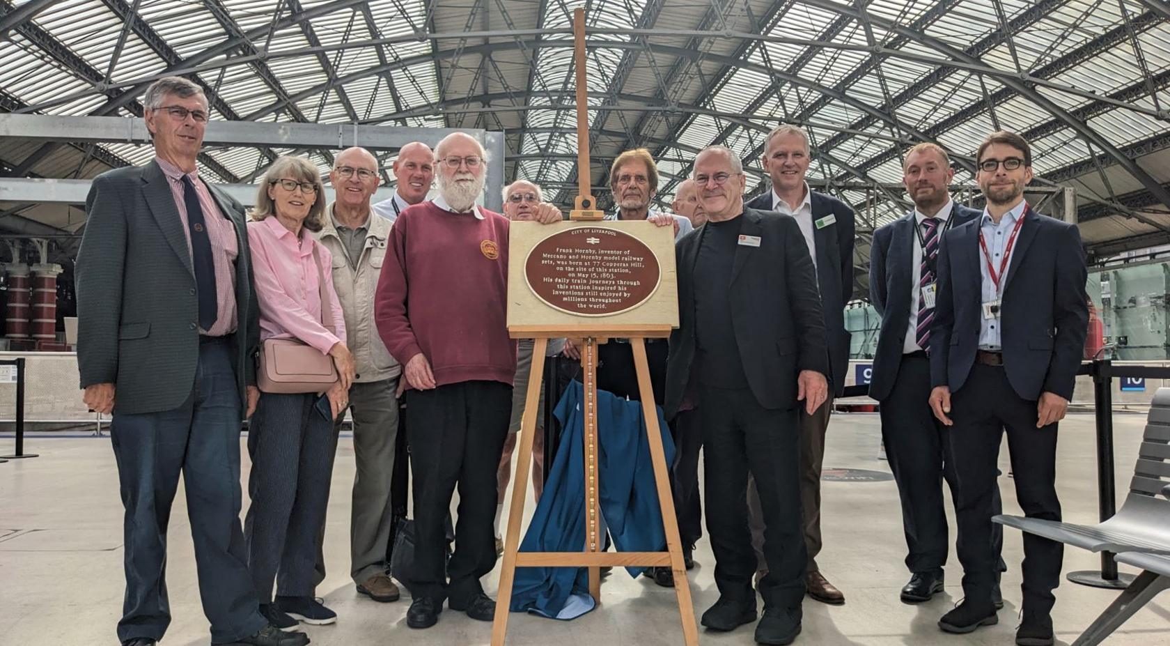 A group of people gather behind a wooden easel bearing a brown plaque at Liverpool Lime Street station.