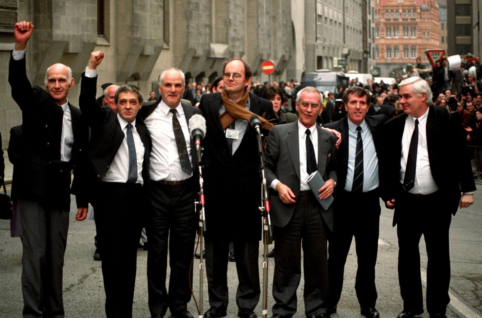 Seven men stand in a line on a busy street. They are all wearing dark suits and ties. The two men on the left are holding their fists in the air in celebration. There are two microphones in front of the men and a crowd of people behind them.