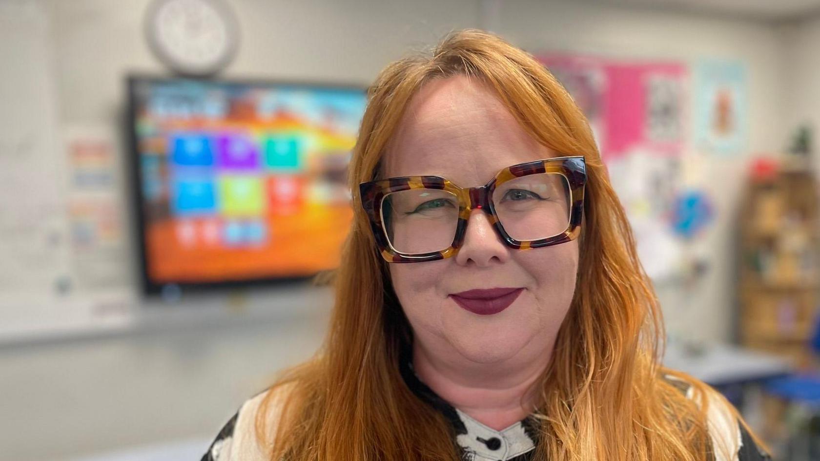 Rhian Evans, an English teacher with long red hair and tortoiseshell glasses smiling at the camera in a classroom