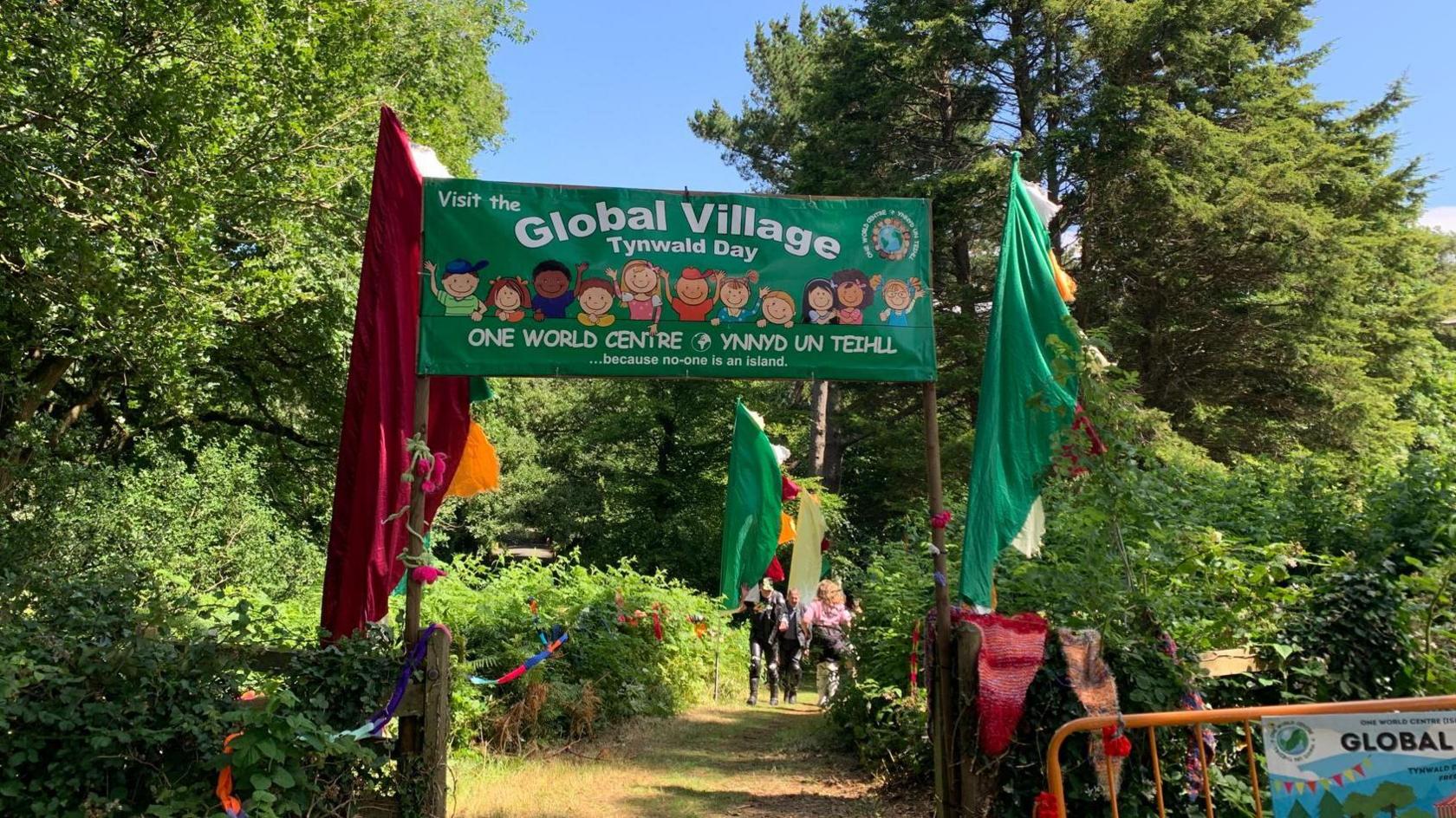 The Global Village green sign makes an archway ahead of a path lined with trees down to the arboretum festivities.