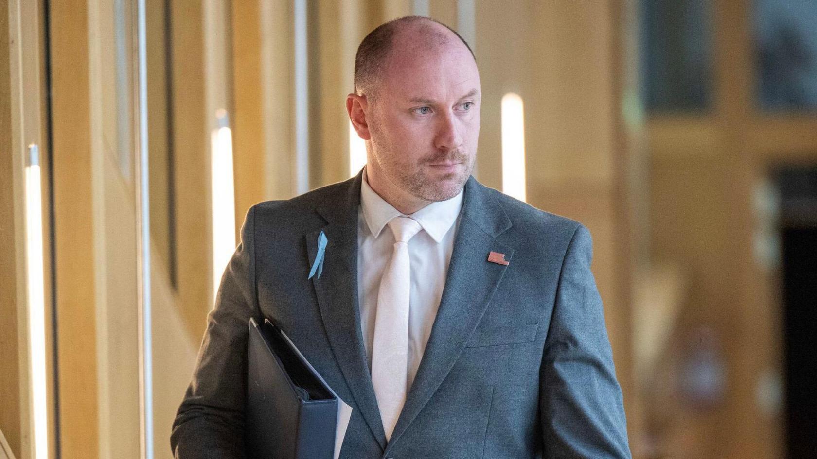 Neil Gray, a bald man wearing a grey suit and white tie, walks down the corridor to the chamber at the Scottish Parliament. He holds a black folder under his arm.