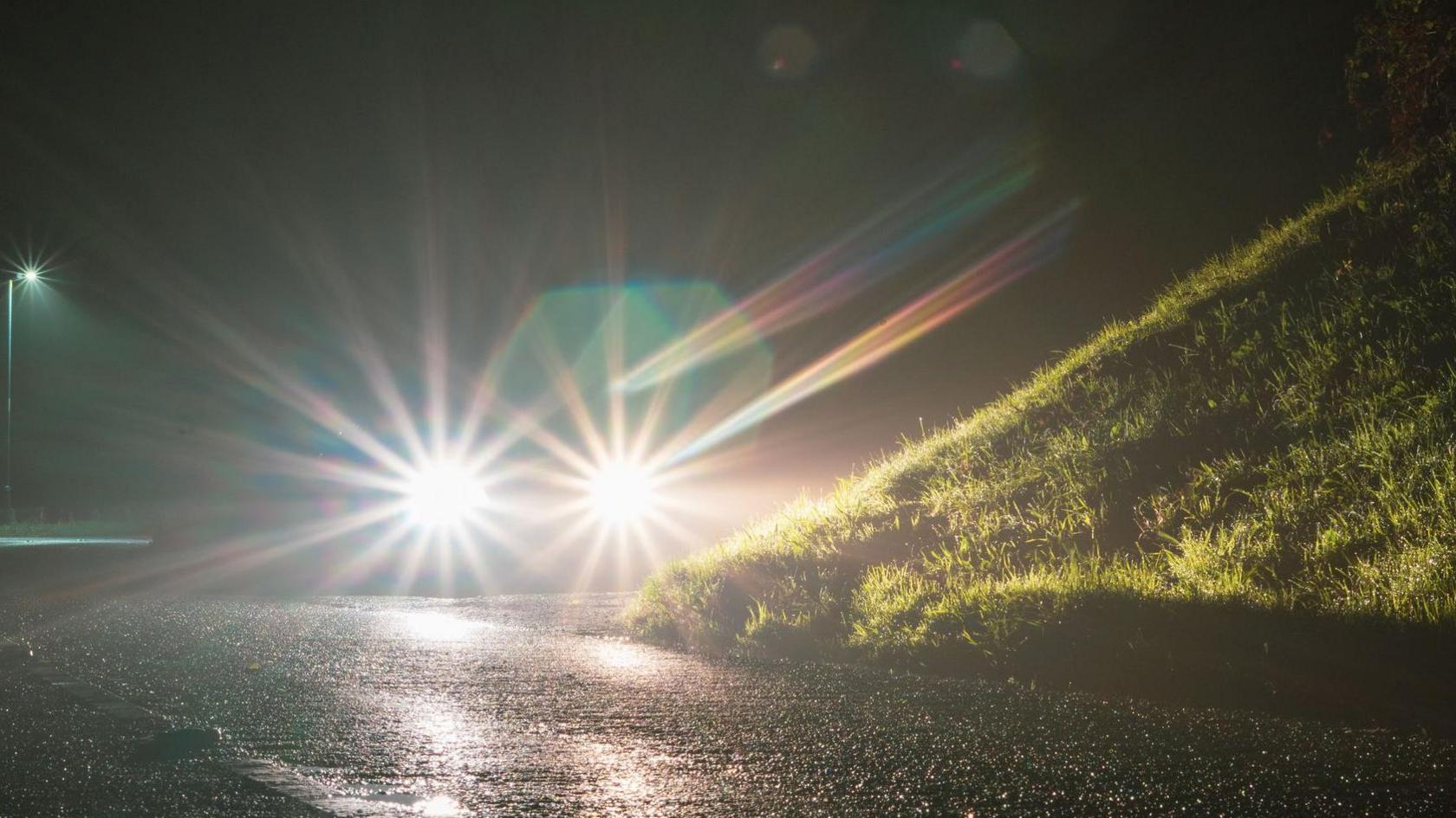 A generic picture showing a car's headlights glowing at night on an empty country road, with a grassy bank on the right.