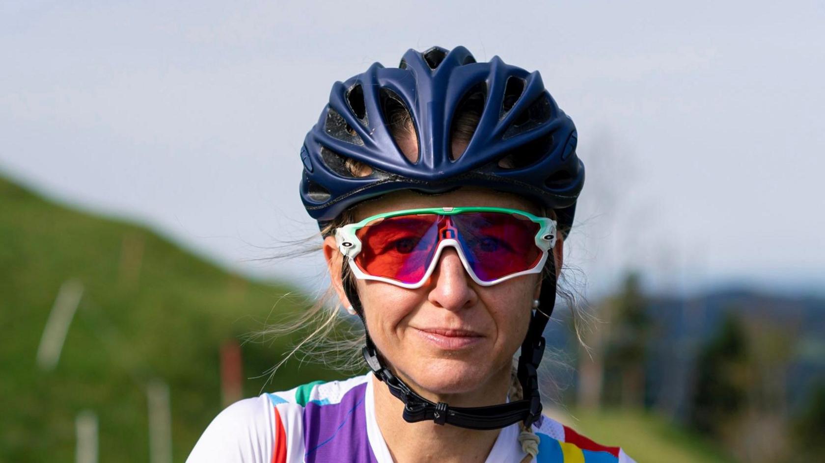 Emma Pooley is looking at the camera and is wearing a blue cycling helmet and glasses. She has long, blonde hair