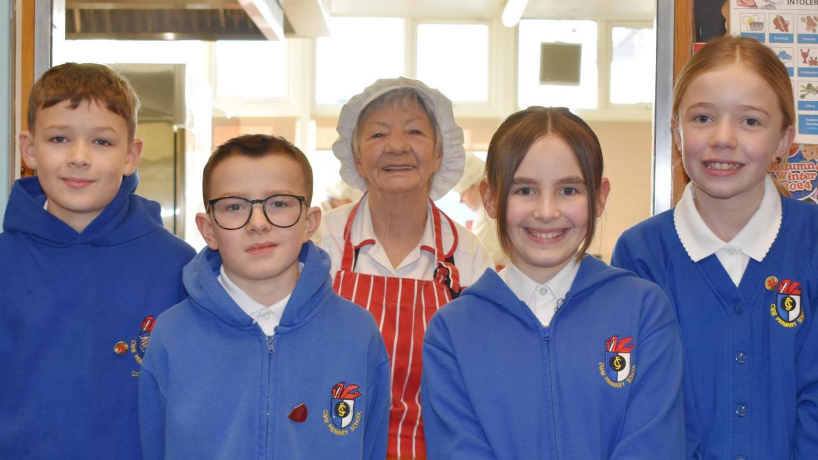 Sonia Blachard in catering hat and white uniform with red and white striped apron on top. She is flanked by four pupils in blue uniform hoodies or cardigans and white polo shirts.