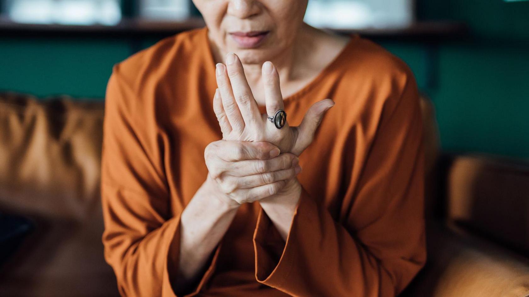 A woman is seen holding her hand in pain. She is wearing an orange top. 