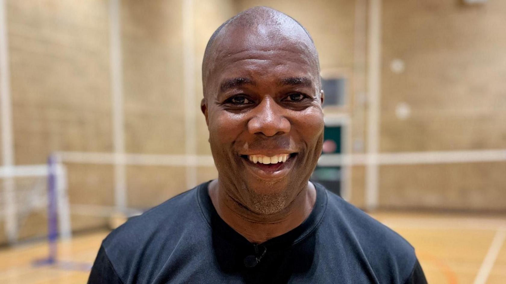 Patrick Lawrence is in the middle of the image standing on a badminton court smiling. He is wearing a navy blue T-shirt. 