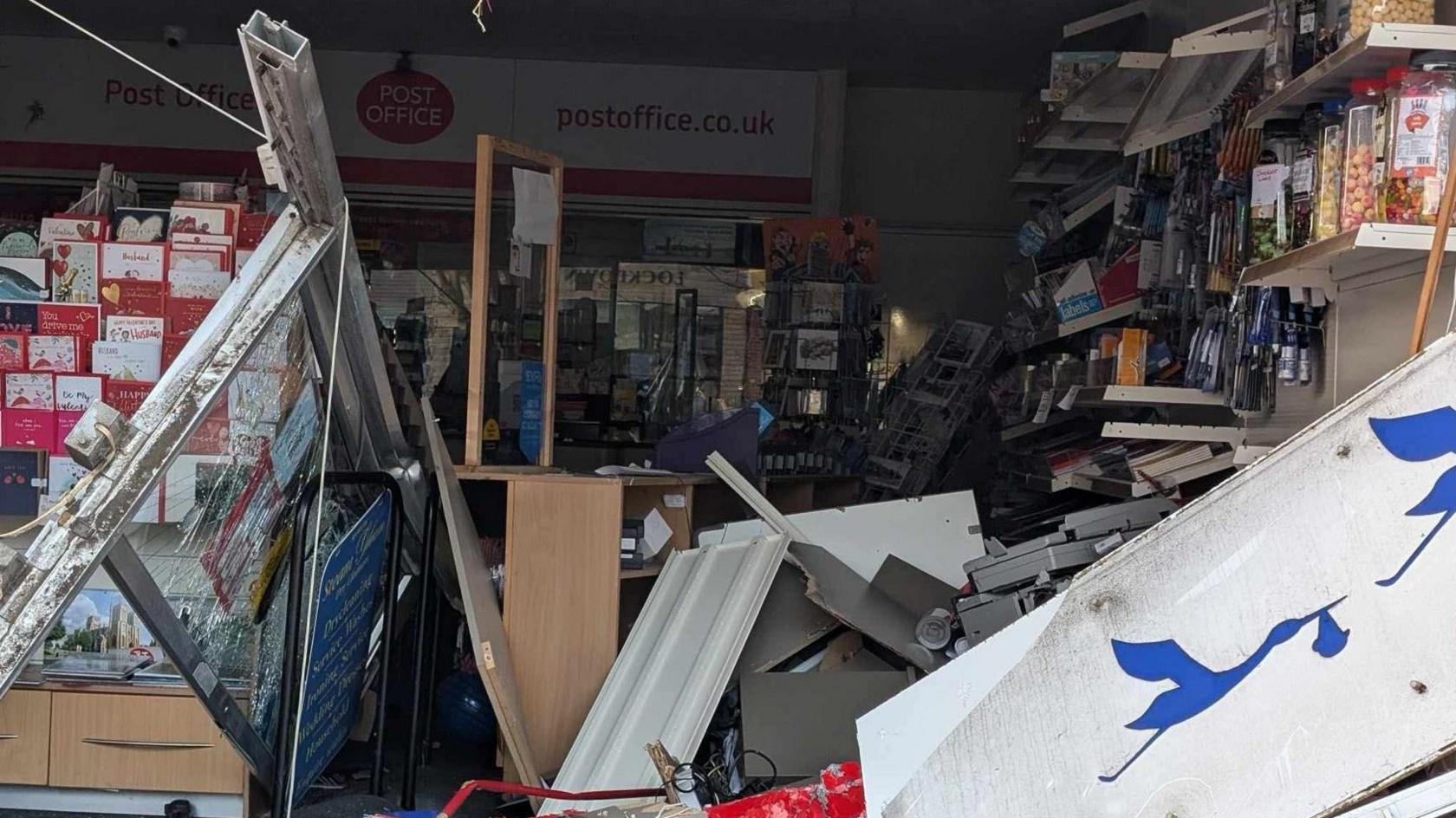 Close up image showing the damage to the post office in Winterbourne. A stand of greeting cards can be seen to the left of the frame and there is a lot of damaged stock and fixtures from the inside of the post office on the floor, blocking the aisle.