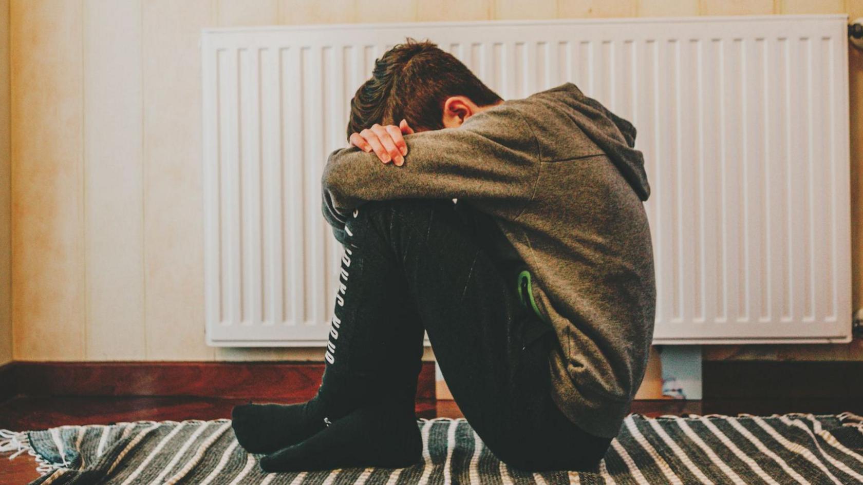 An upset-looking young boy wearing a grey hoodie sits hunched over on the floor with his head tucked into his arms, which are folded on top of his raised knees