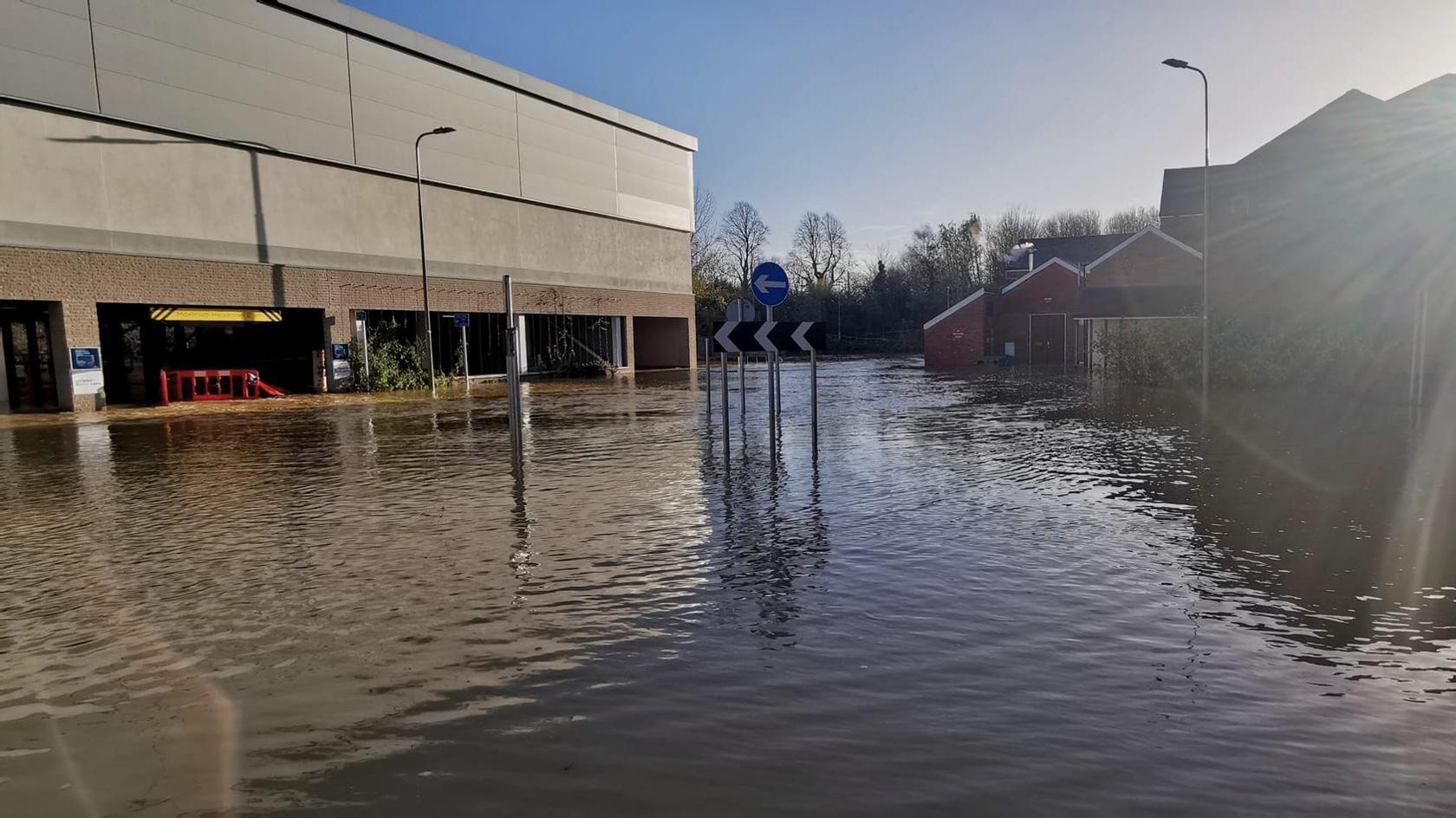 A flooded street with a grey multi-storey carpark next to it