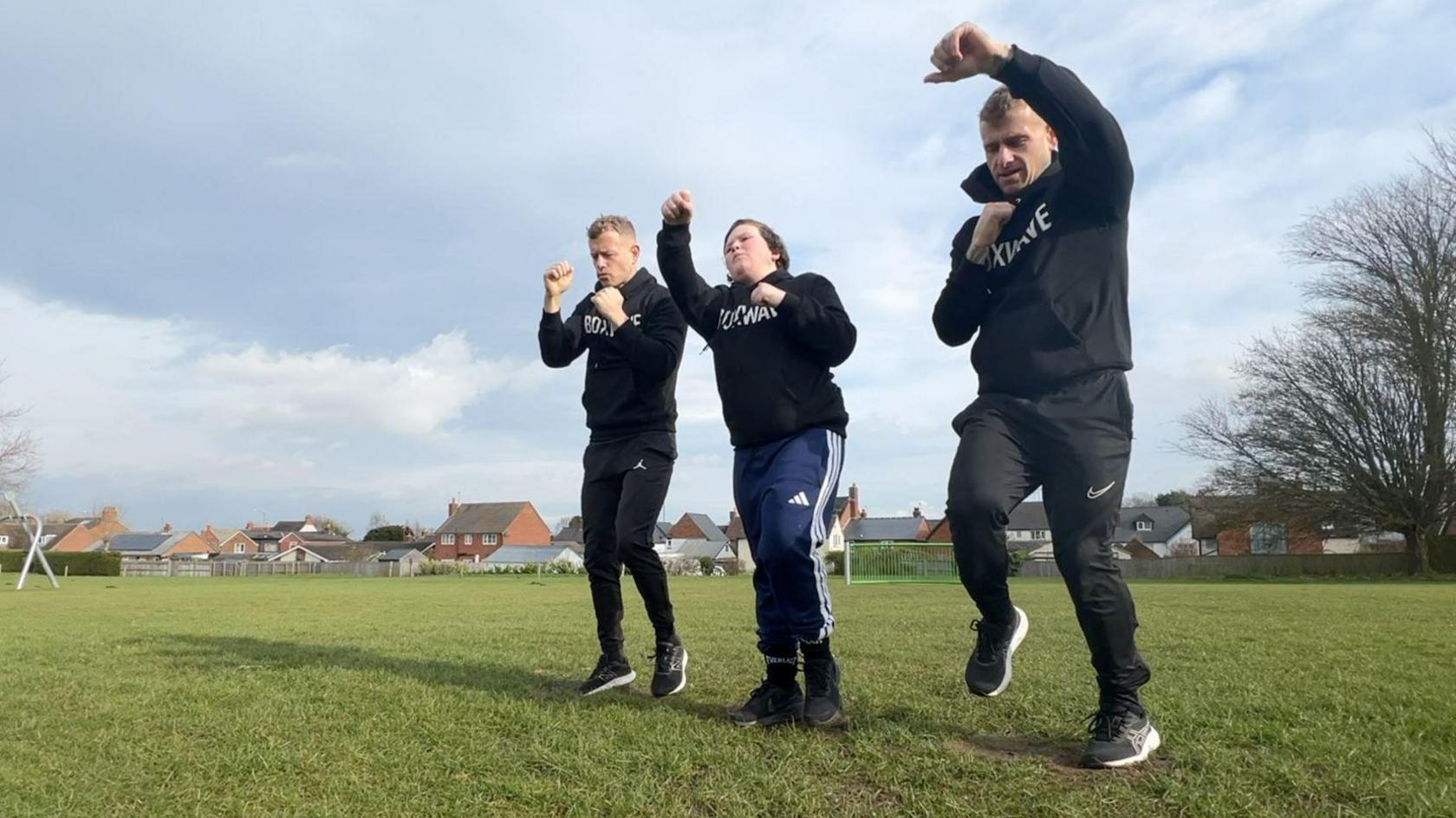 Three people wearing black jumpers taking part in fitness training on grass.