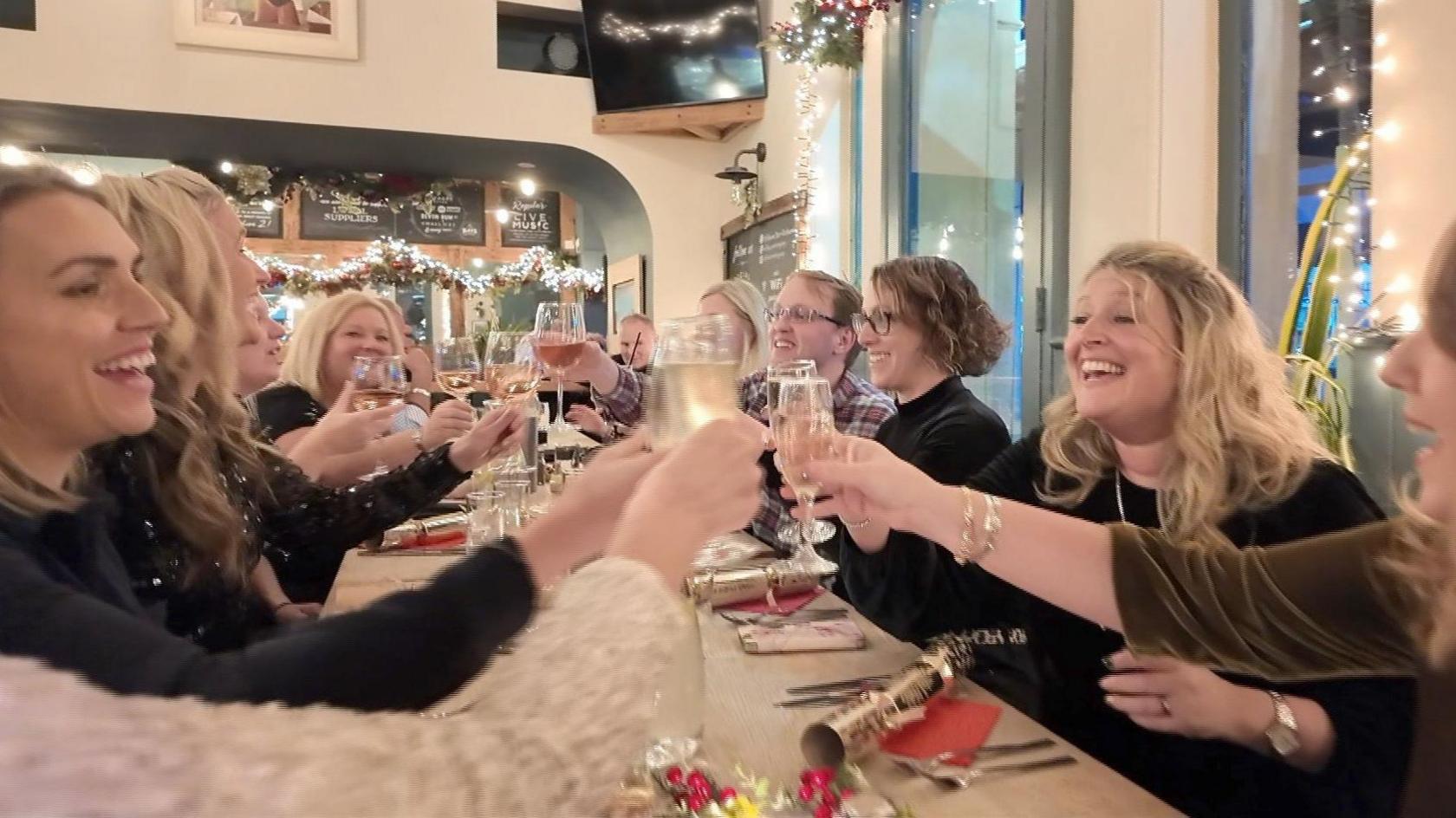 A group of people sit around a long dining table in a bar. There are Christmas crackers on the table and Christmas decorations in the background. The group are holding their wine glasses up in a 'cheers' action. 