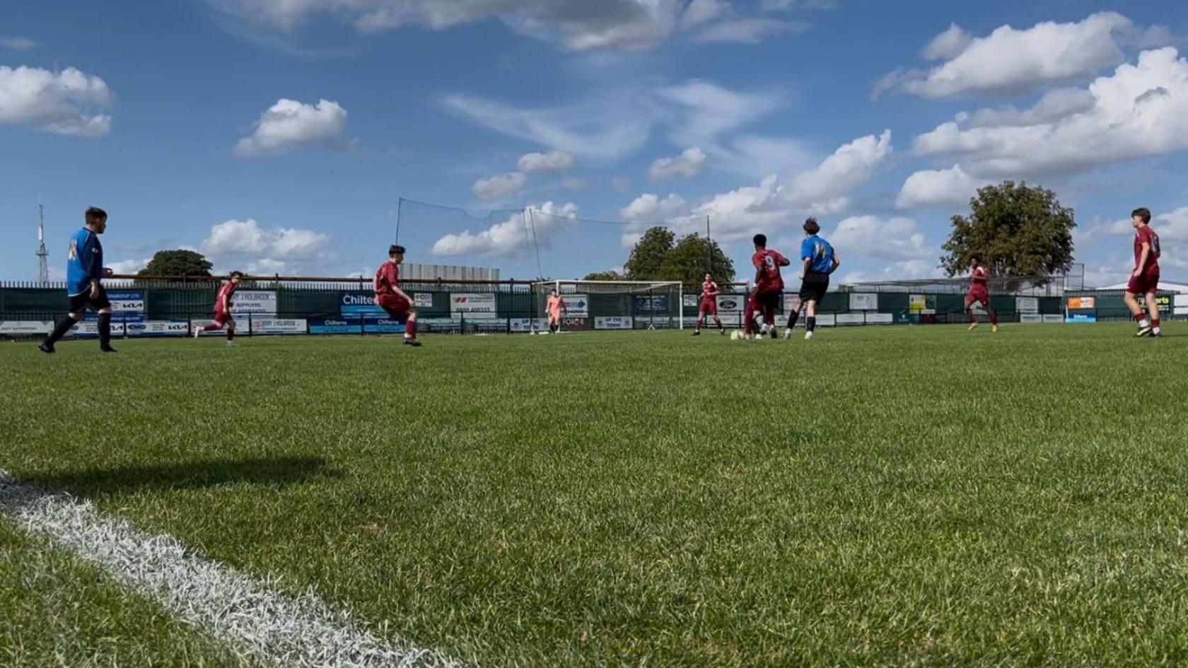 A match between Thetford Town Football Club and Thetford Bulldogs' under 18s. There are nine players on the pitch wearing either bright blue or maroon strip.