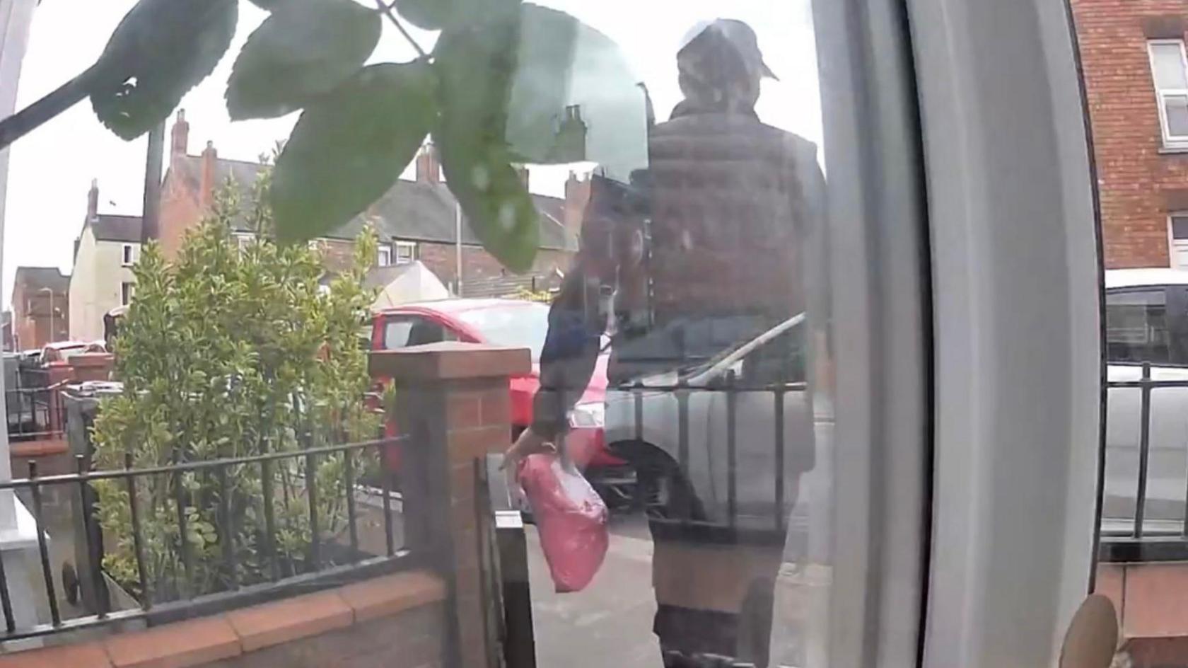 Doorbell camera footage of a man dressed in black with a black cap walking out of a front garden with a pink parcel.