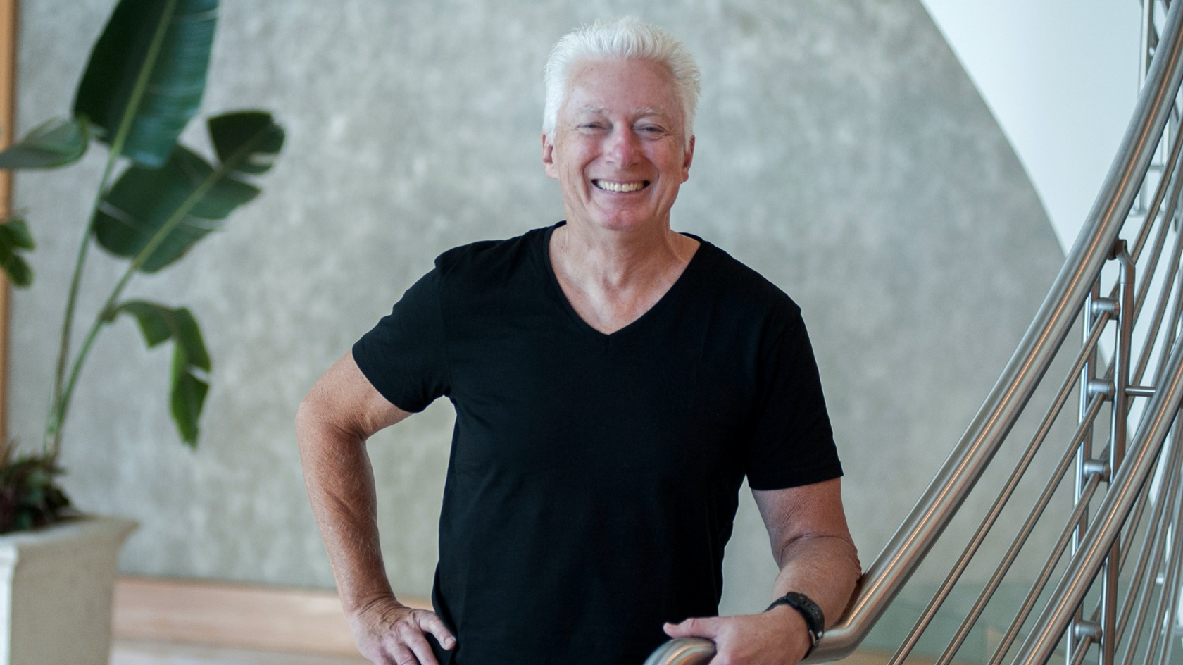 Former P&G CEO Alan Lafley poses in a black t-shirt whilst standing at the bottom of a stair case with a silver metal railing 