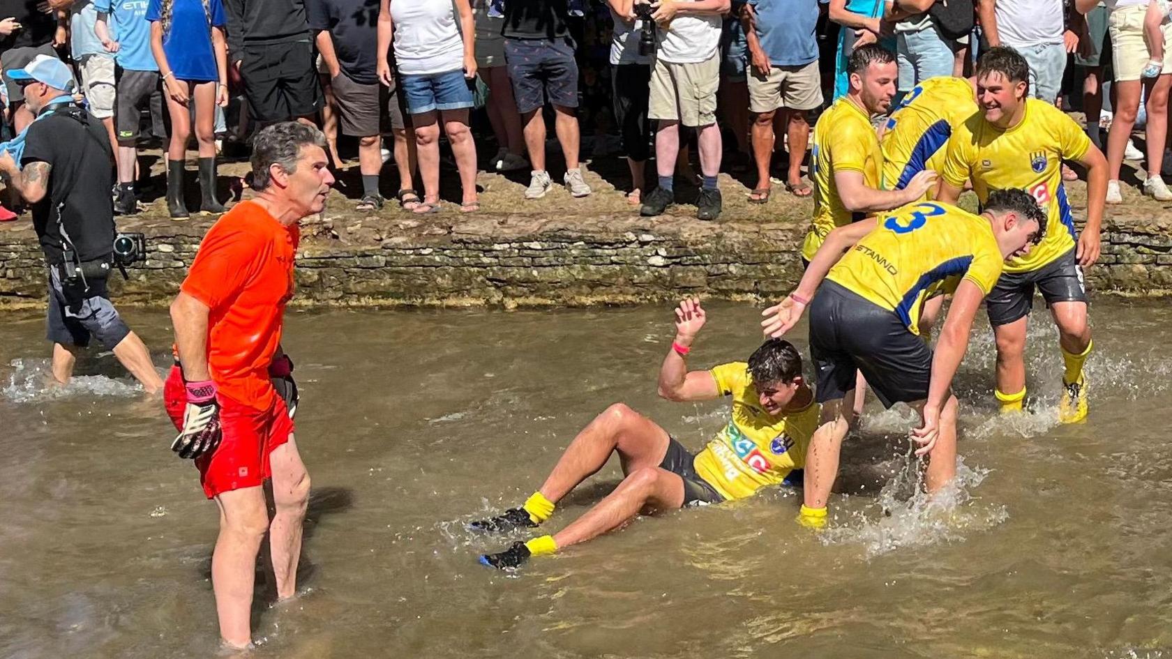 A group of players in yellow and blue sports in a shallow river. Spectators line the grassy riverbank, some seated and others standing, closely watching the match.