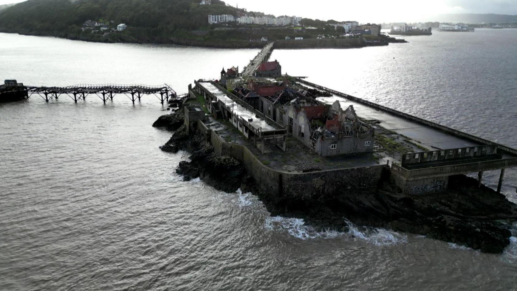 Drone shot of old pier that has a building that is in disrepair. The roof is off. The waves are hitting the island and there's a view looking back into the seaside town of Weston-super-Mare.