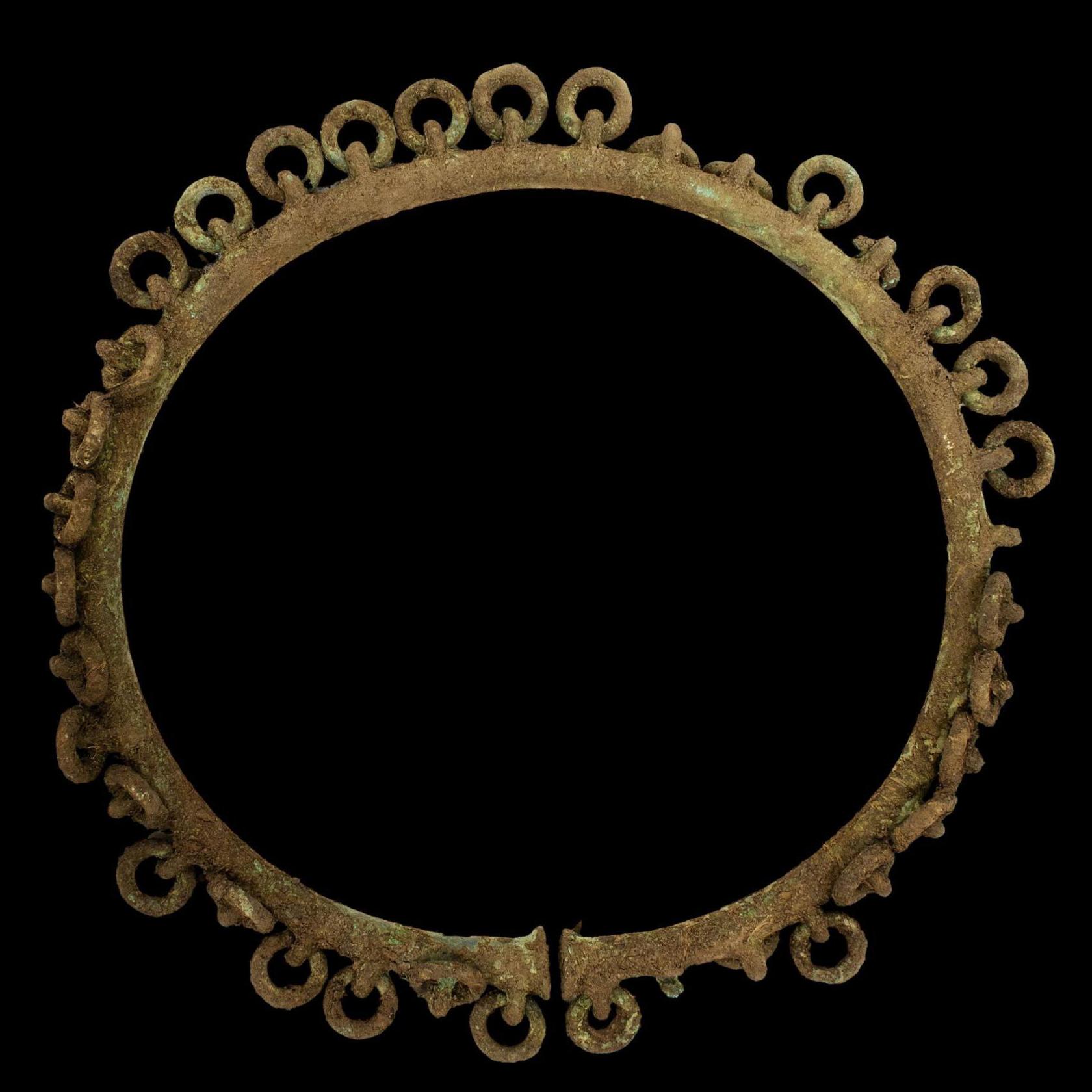 The penannular ringed ornament has been photographed against a black background. It is a circle of bronze, but with a small opening. Smaller rings are attached around the edge of the circular object.