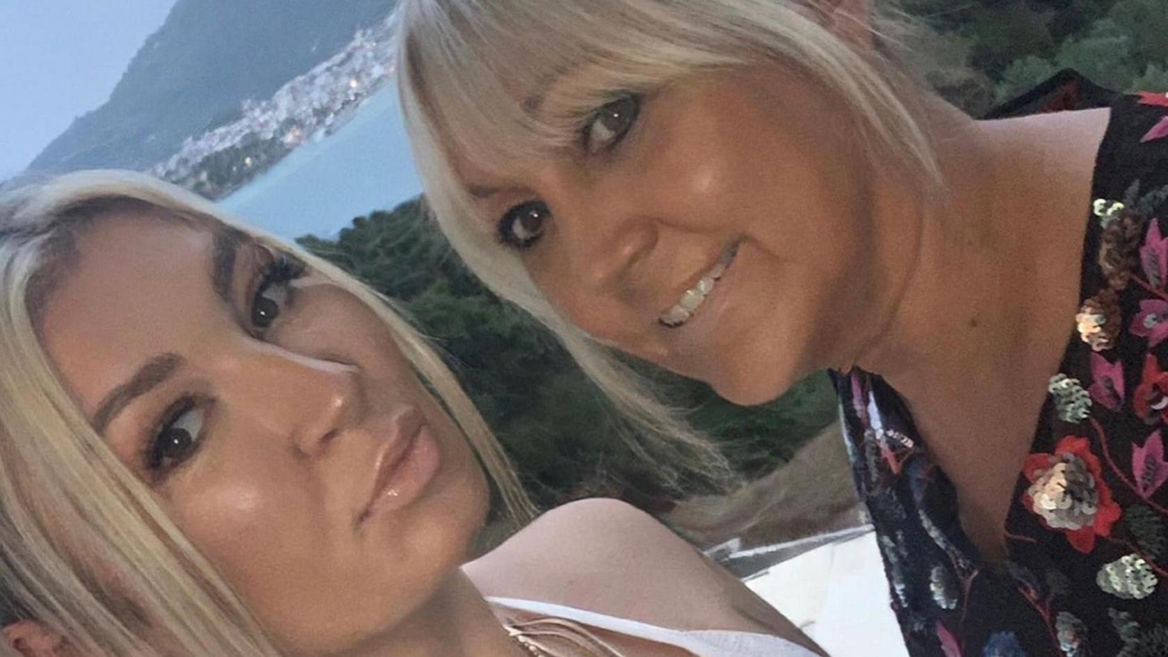 A mother and her daughter pose for a selfie overlooking the sea. Both have long blonde hair tied back. The young woman on the left is pouting and wearing a strapless white top. Her mother has a black top with silver sequins and red and purple flowers.
