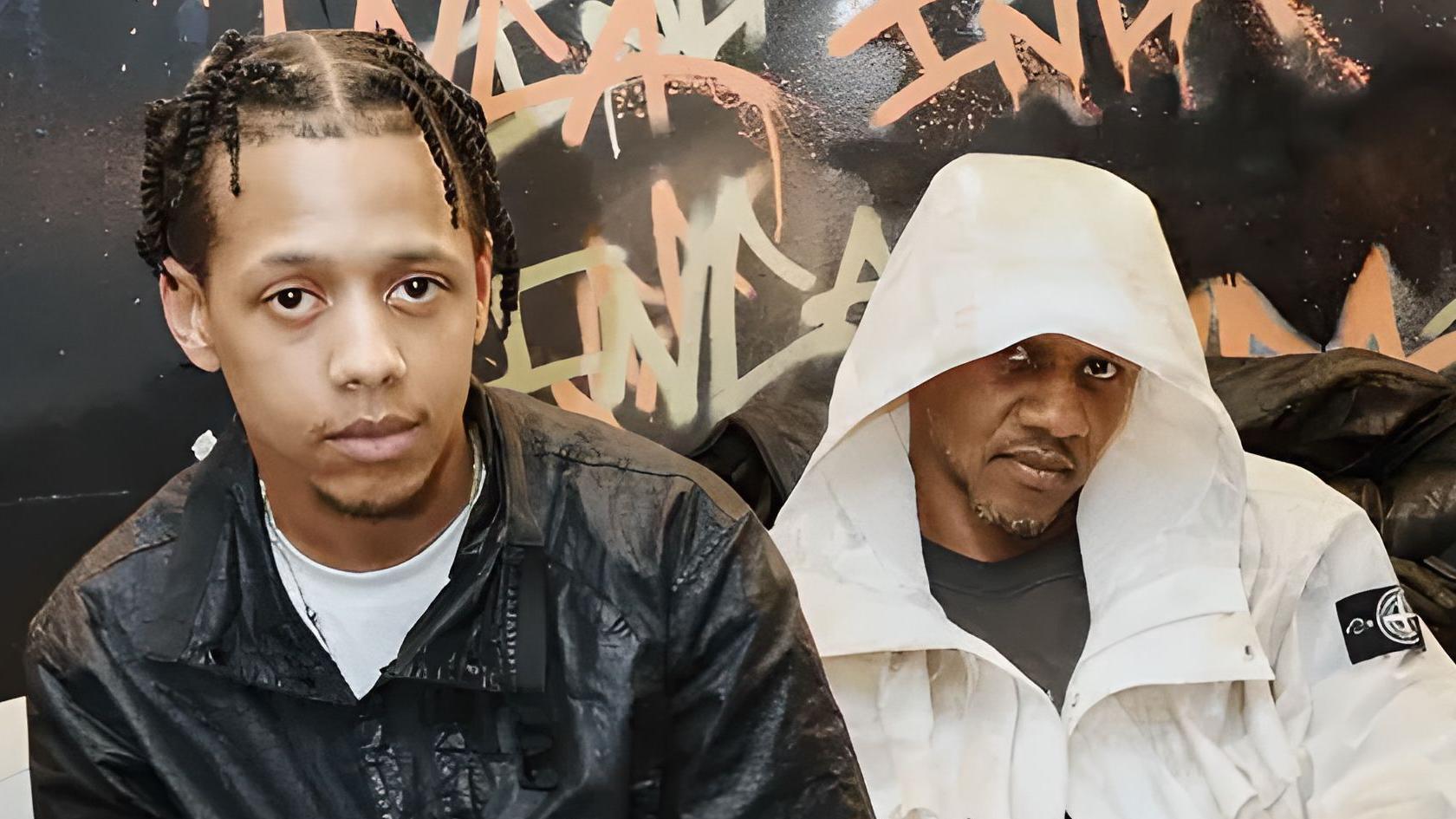 ML and Giggs sit on a sofa, ML leaning forward in front of his dad. ML wears a dark short sleeved shirt while Giggs wears a long sleeved white jacket with the hood up. They're pictured in front of a dark wall that's been graffitied.