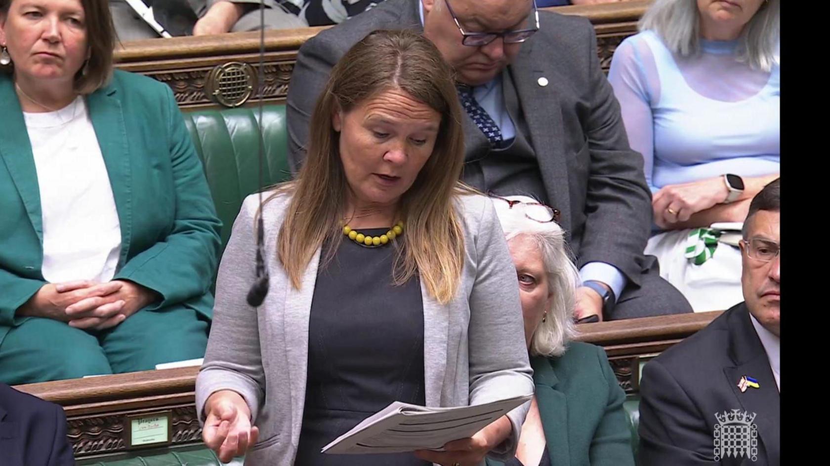 Vikki Slade MP is speaking about Axel's case from the opposition benches in Parliament. She is wearing a black top, grey jacket and a yellow necklace.