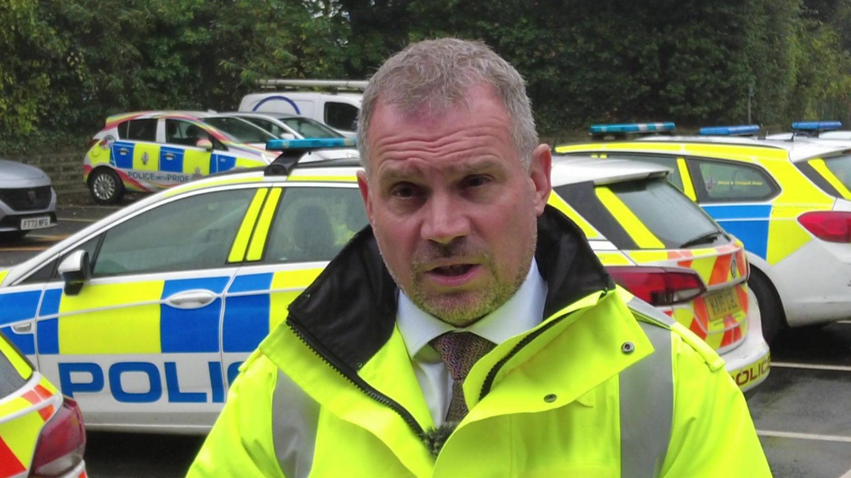 A police officer wearing a high-vis yellow jacket in front of several police cars at a police station.