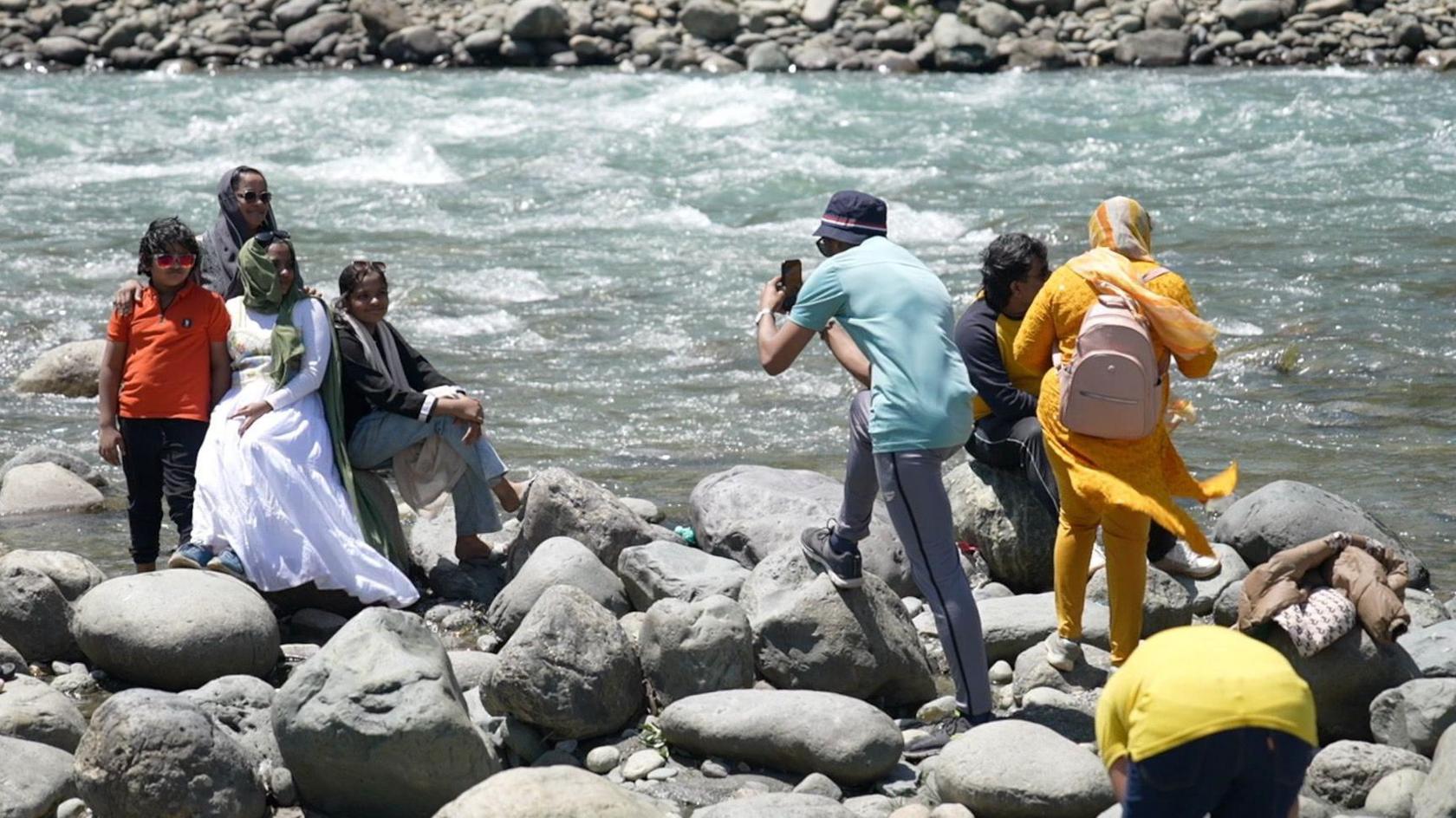 The image shows tourists sitting on the rocks by a river in Pahalgam town, with some families posing for a photograph.