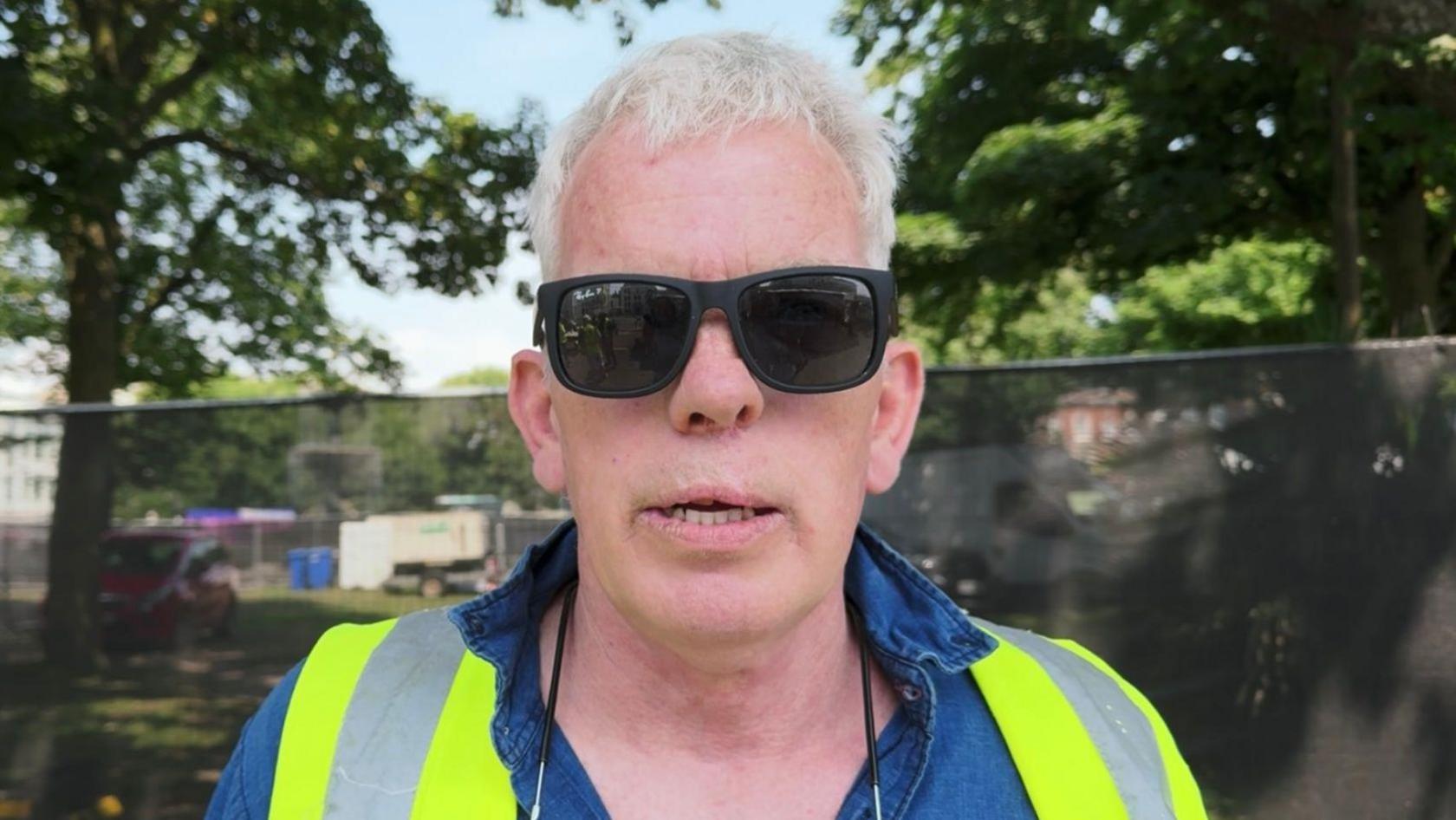 Steven, who is facing the camera, wears black sunglasses, a denim shirt and a sleeveless hi-vis jacket. There are trees and high fencing behind him