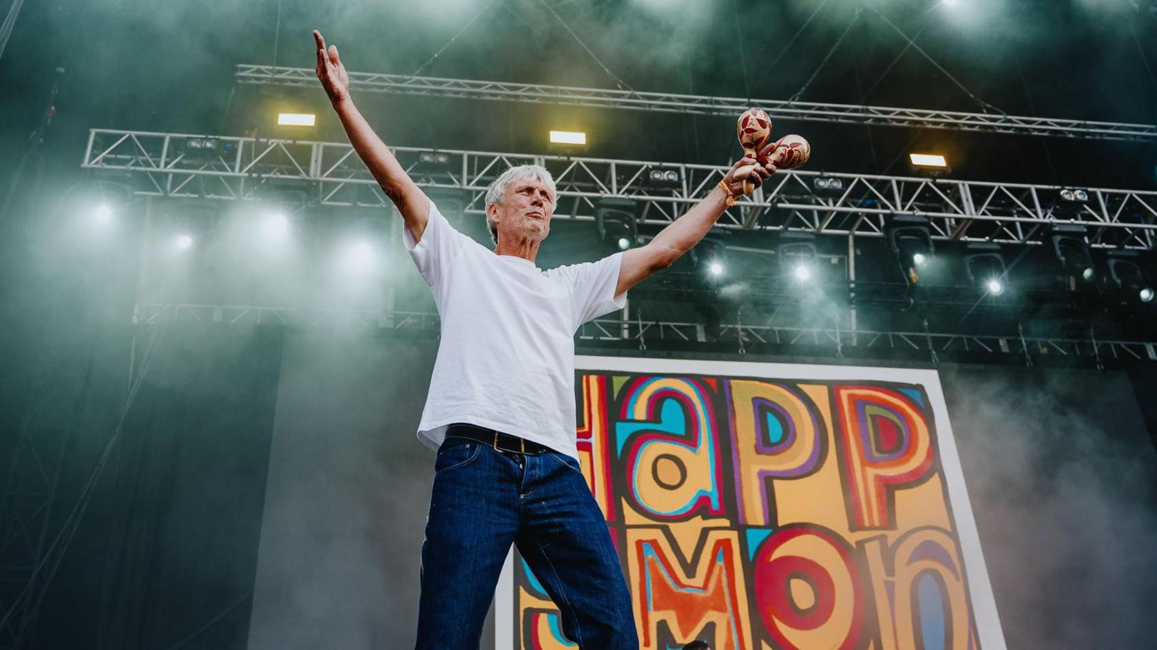 Bez from the band Happy Mondays stands on stage at Forwards Festival in Bristol. He is wearing a white T-shirt and dark blue jeans and is holding maracas in his hand. Behind him is the band's recognisable name in a colourful poster.