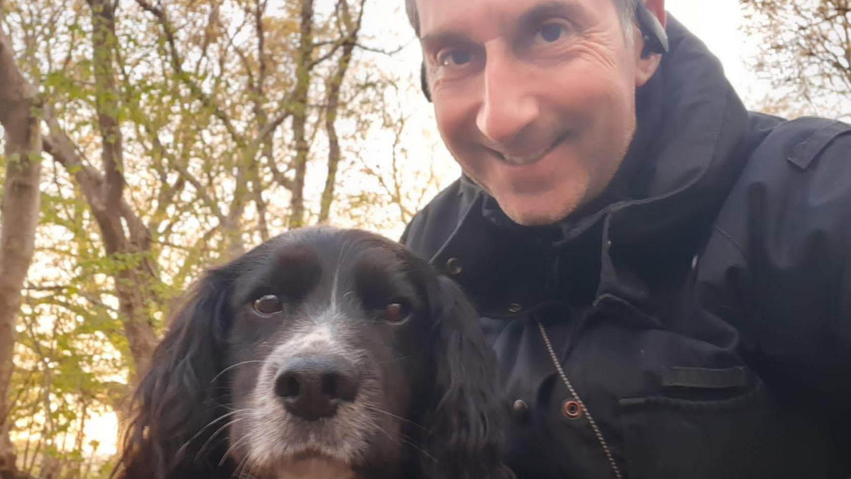 Sherlock looking at the camera lens as his handler Paul Osborne, a man with short hair wearing a dark jacket and earphones, poses for a picture with him in front of some trees