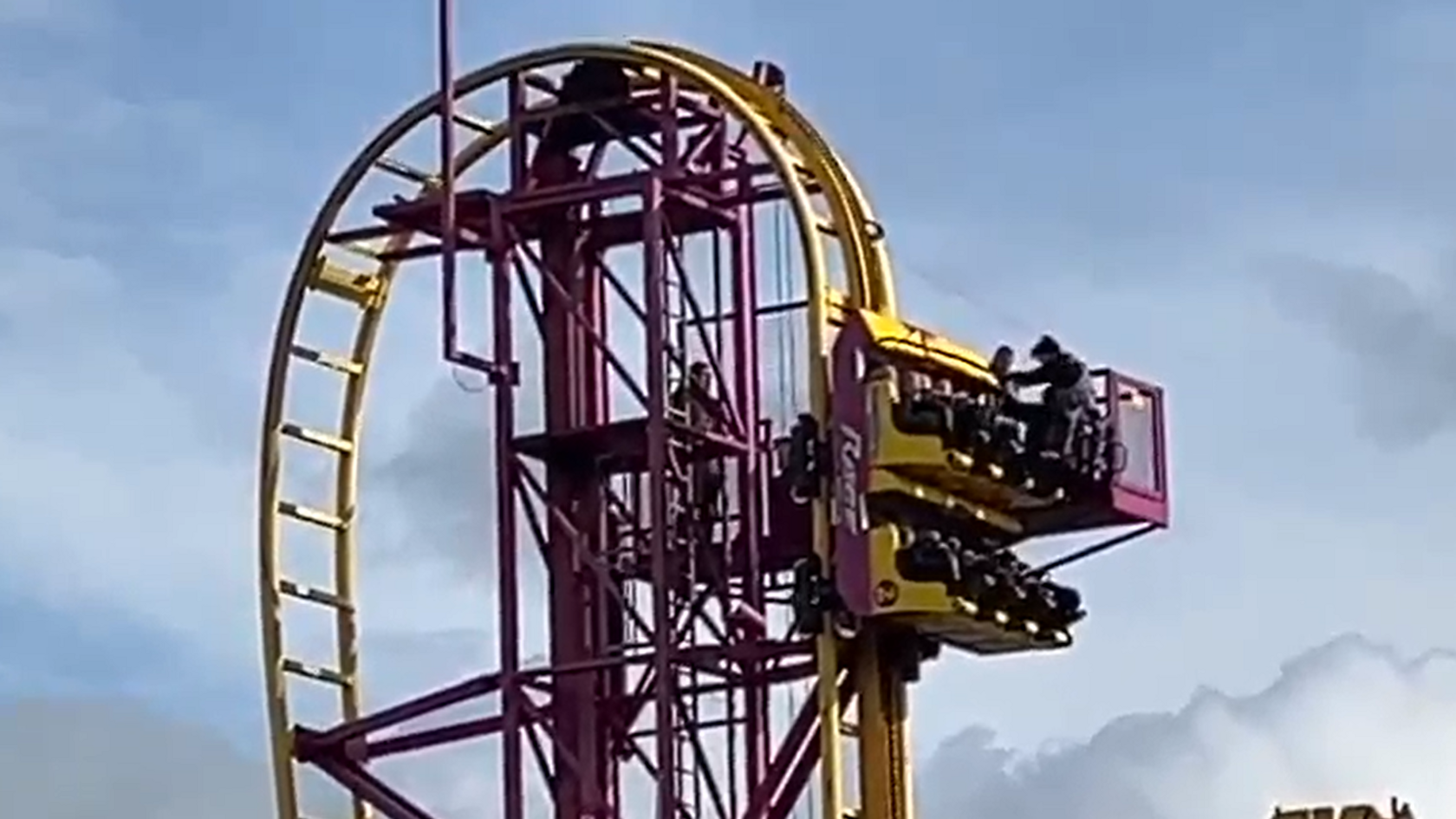 A ride operator stands on a moveable platform next to the roller coaster cart, which is near a high point on the track. The yellow cart, which is facing upwards, has two rows of four black seats. The operator is helping someone out of the cart