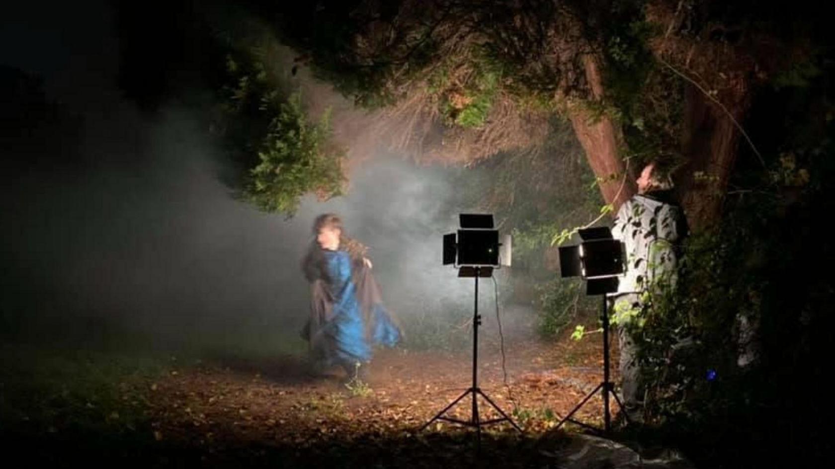 An actress in Victorian clothing running through a dark park. She is illuminated by stage lights and a member of the film crew is standing behind the lights.