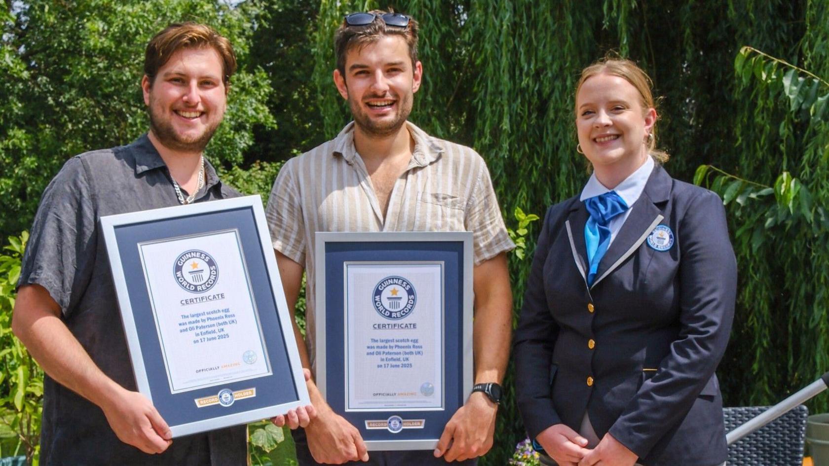 Phoenix, on the left, is stood smiling holding a Guiness World Record certificate, next to Oli, in the middle, who is also smiling and holding a certificate. The female adjudicator, in a blue uniform is stood on the right also smiling. They are outside, with large green trees behind them.