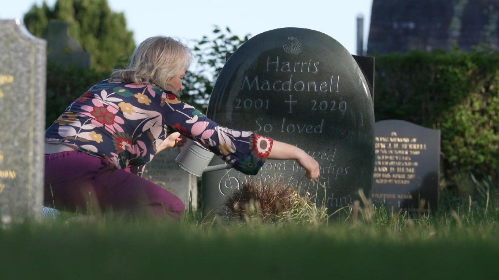 A woman with a flowery blouse crouches with a water in front of a gravestone.