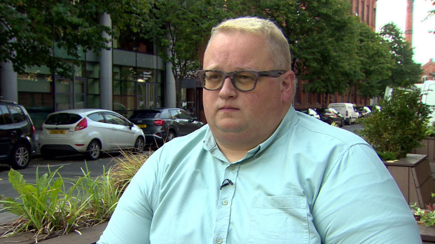 Man with blonde hair, wearing dark rimmed glasses looking stern. He is wearing a light blue shirt. Behind are cars and some green foliage.  