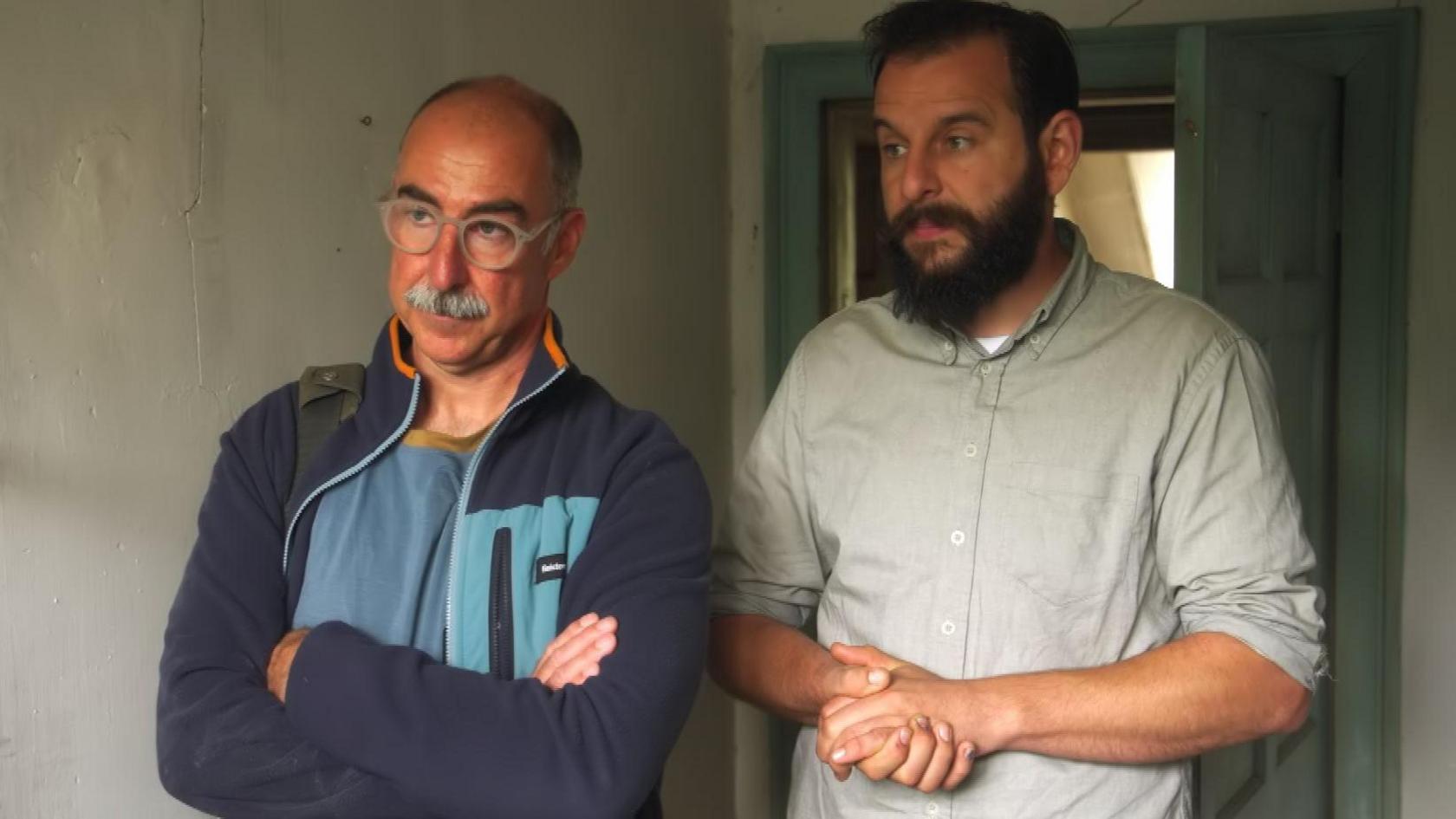 A man with grey hair and a moustache, he is standing with his arms crossed. Next to him is a man in a khaki shirt with dark hair and a beard is is also looking at the plaster.