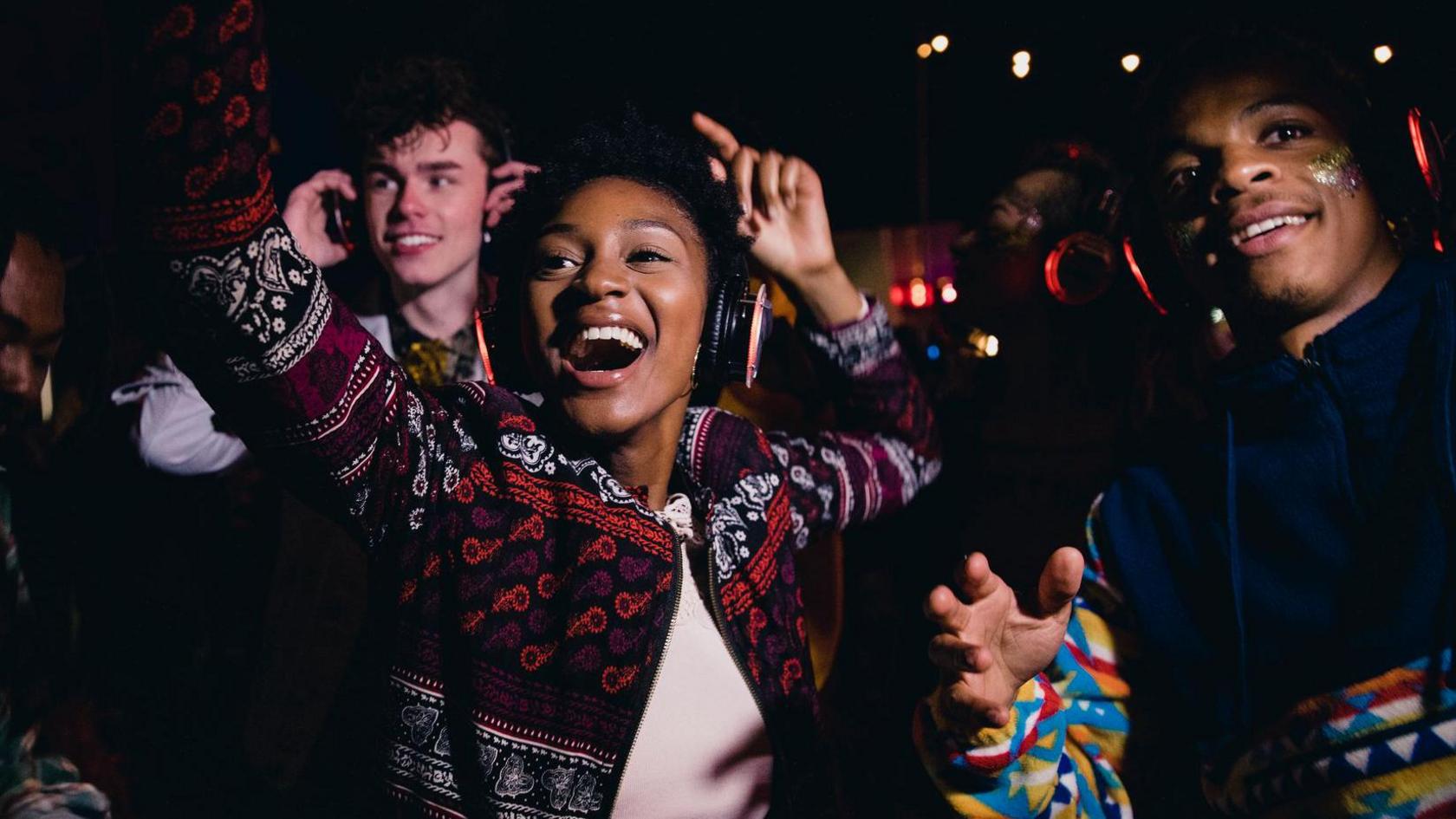 Young people dance and raise their arms in the air while wearing headphones, in a dark place with a trace of some lights in the background