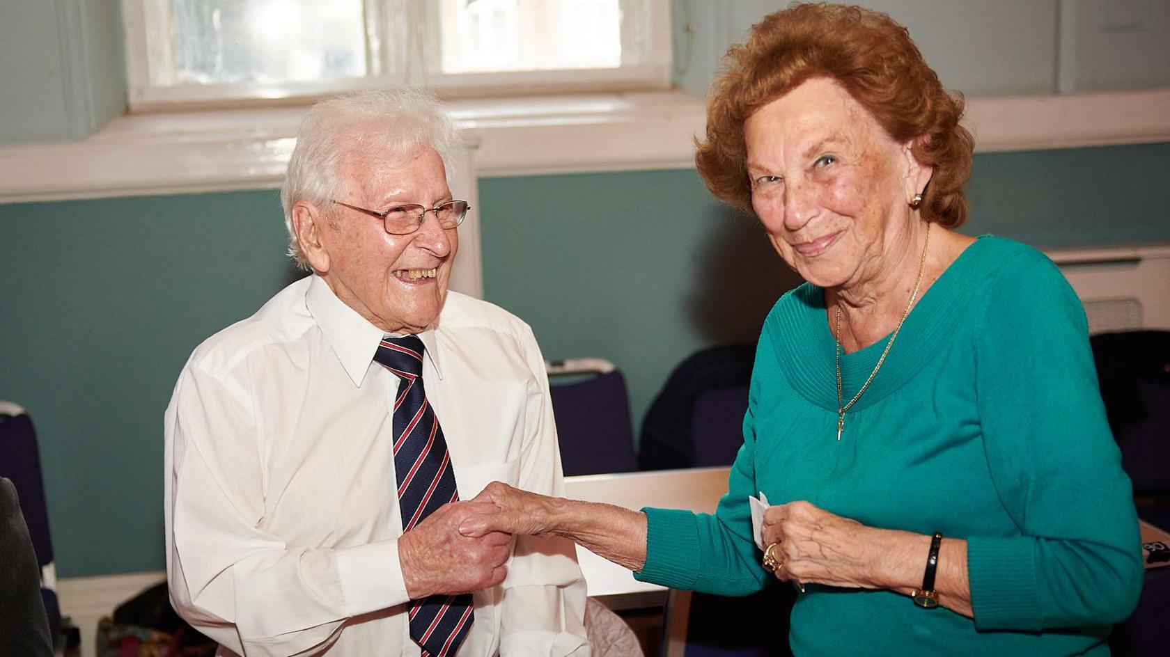 Mr Bickley, in white shirt and striped tie, holds a woman's hand. She has auburn hair and a green top with a thin gold necklace and a watch on her wrist. She slightly smiles at the camera while Mr Bickley looks at her