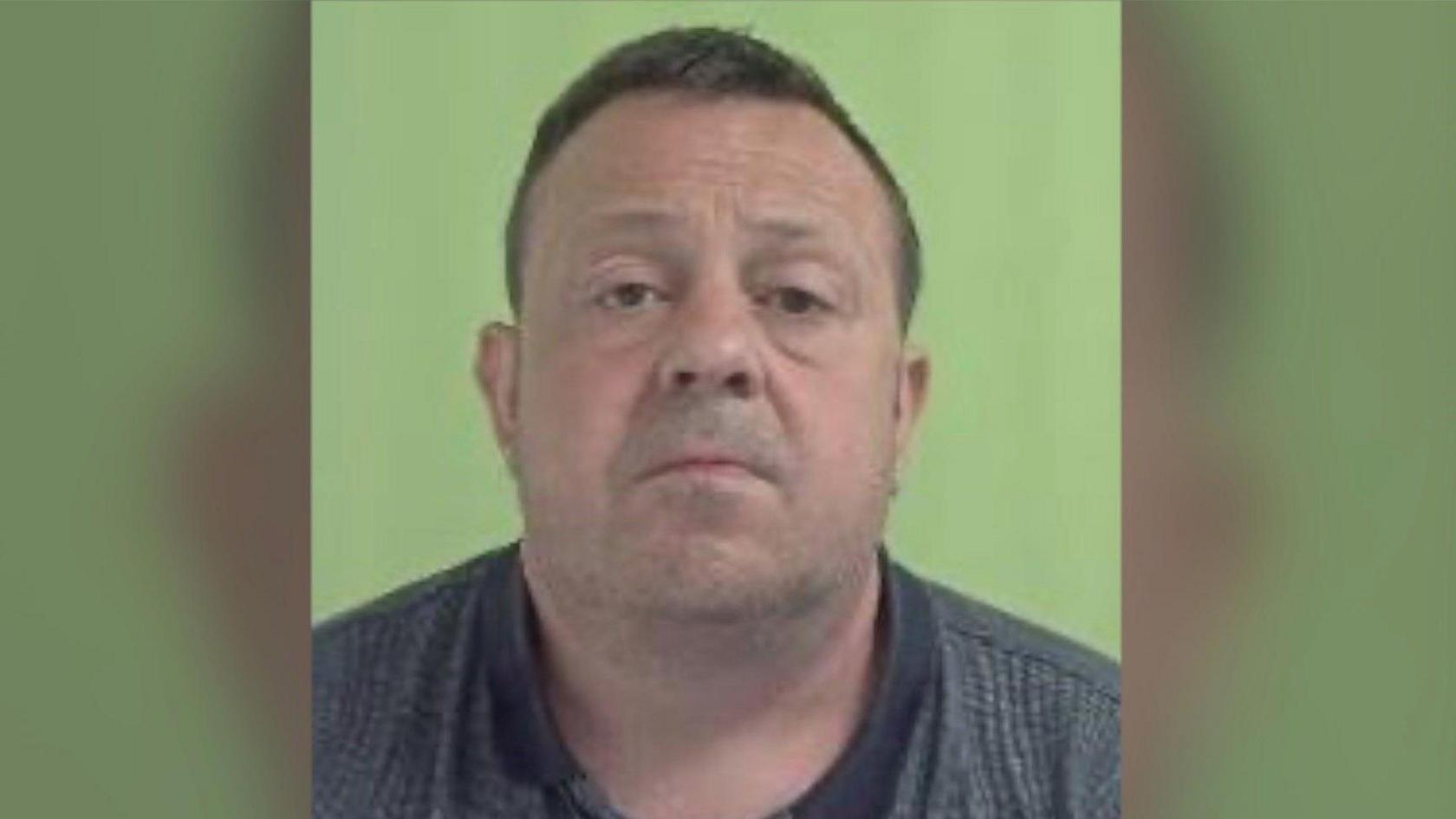 A police mugshot of a middle-aged man in a grey polo shirt, he has short brown hair 