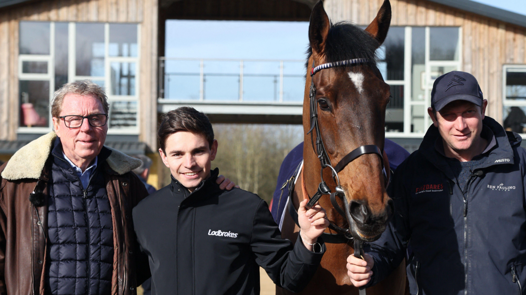 Race horse owner Harry Redknapp, trainer Ben Pauling and Jockey Ben Jones pose with Cheltenham Festival hopeful The Jukebox Man at the Ben Pauling Stables. There is a wood-panelled building behind them which is split in two and connected by a glass balcony walkway. It is a sunny day.