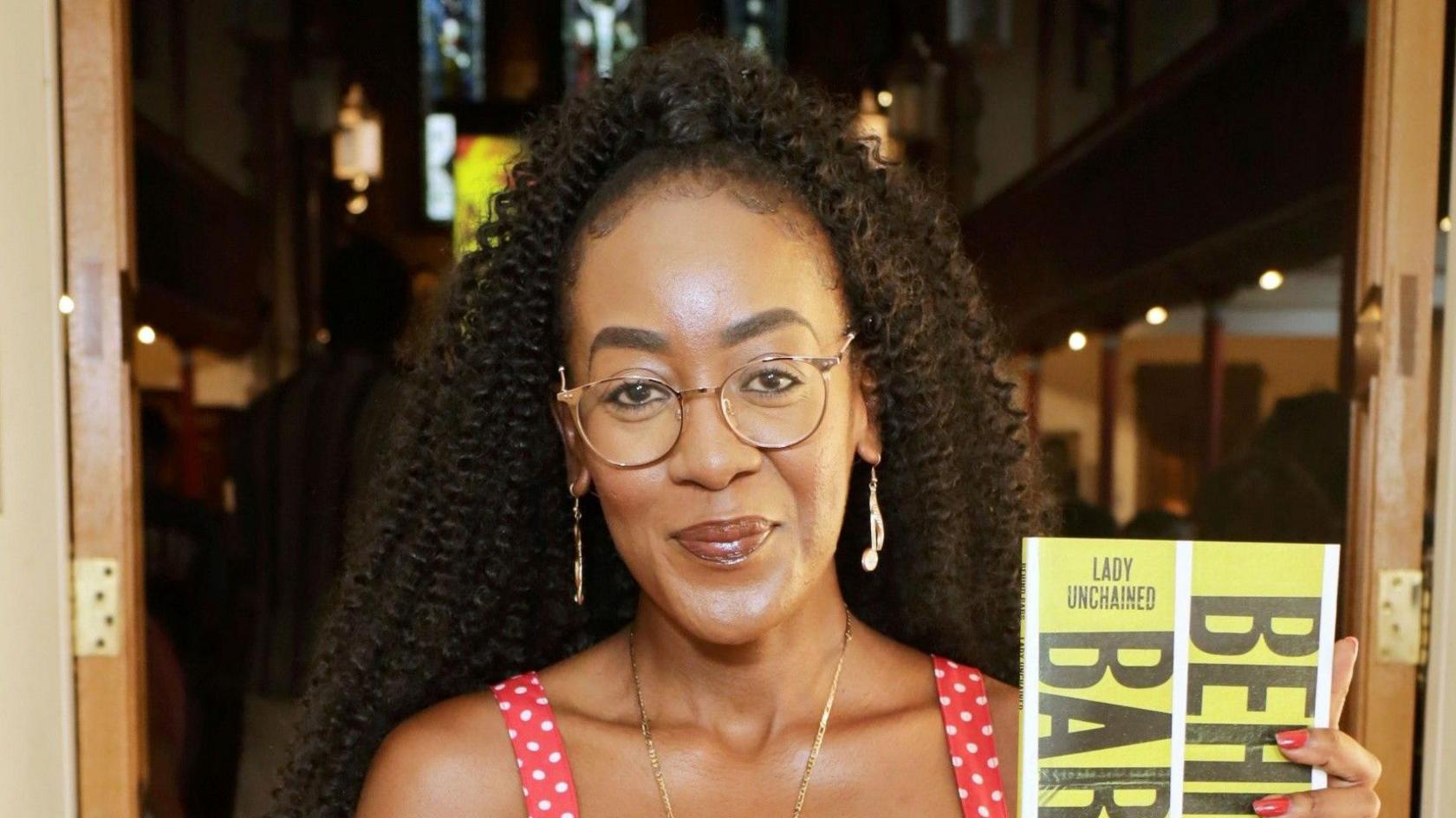 A woman wearing a red and white polka dot dress standing in a doorway holding a yellow book which reads "Behind Bars". She is half-smiling, wearing glasses and long earrings.