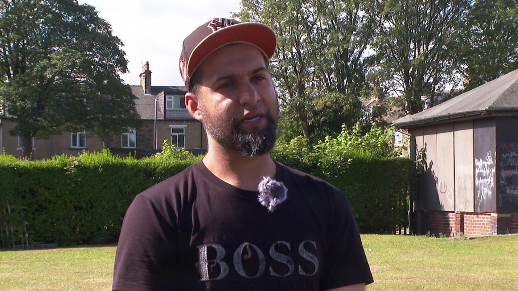 A young man wearing a baseball cap and a dark t-shirt featuring the word 'Boss' stands in a public park. A hedge, small building and trees can be seen behind him.
